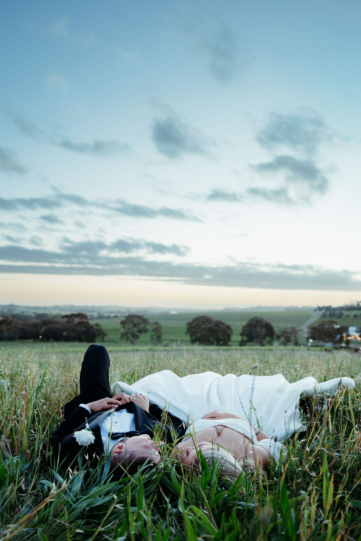 Bride and groom lying in a lush green field at dusk, sharing a romantic moment under a soft blue sky.