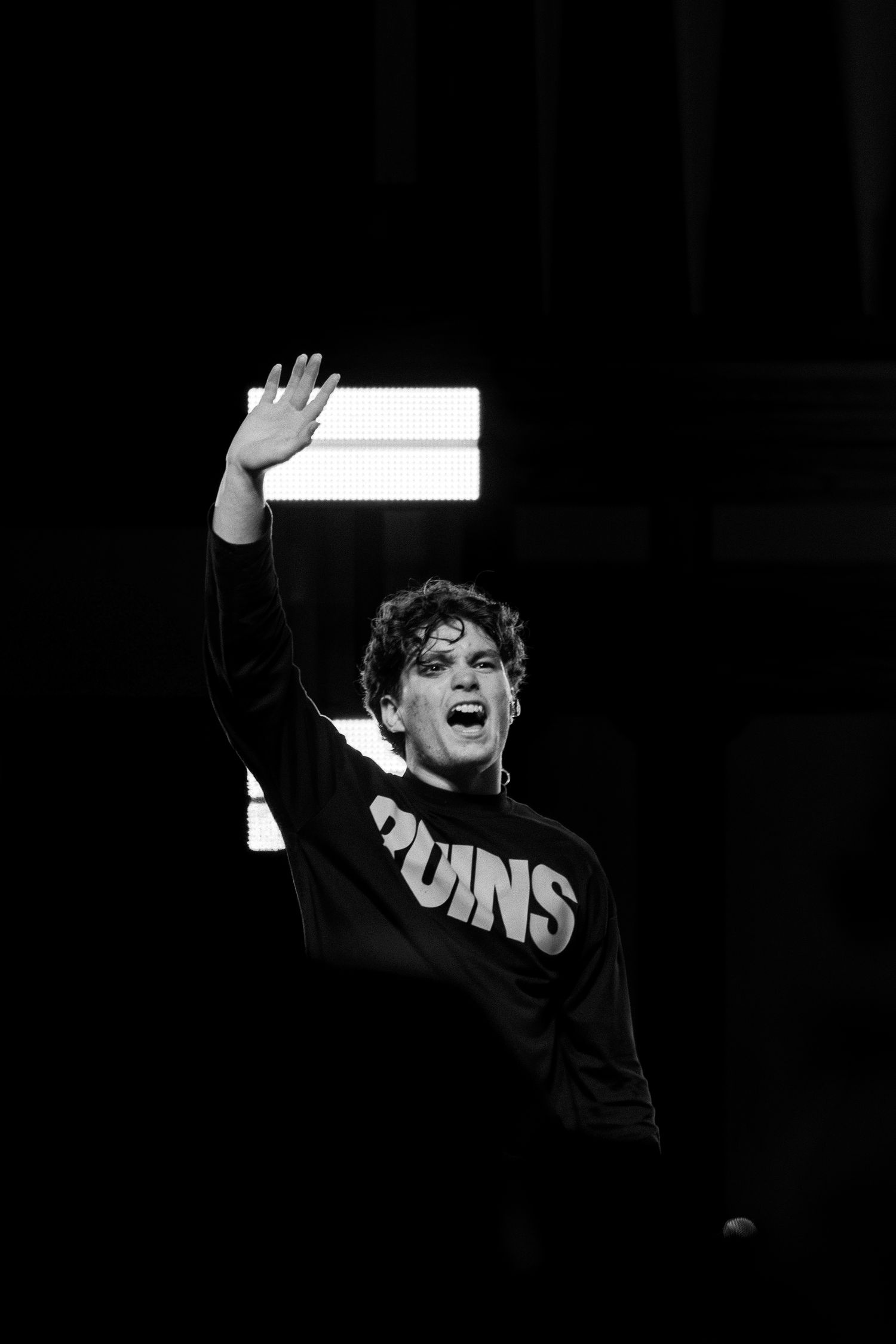 Black and white photo of a young performer in a Ruins sweatshirt waving on stage.