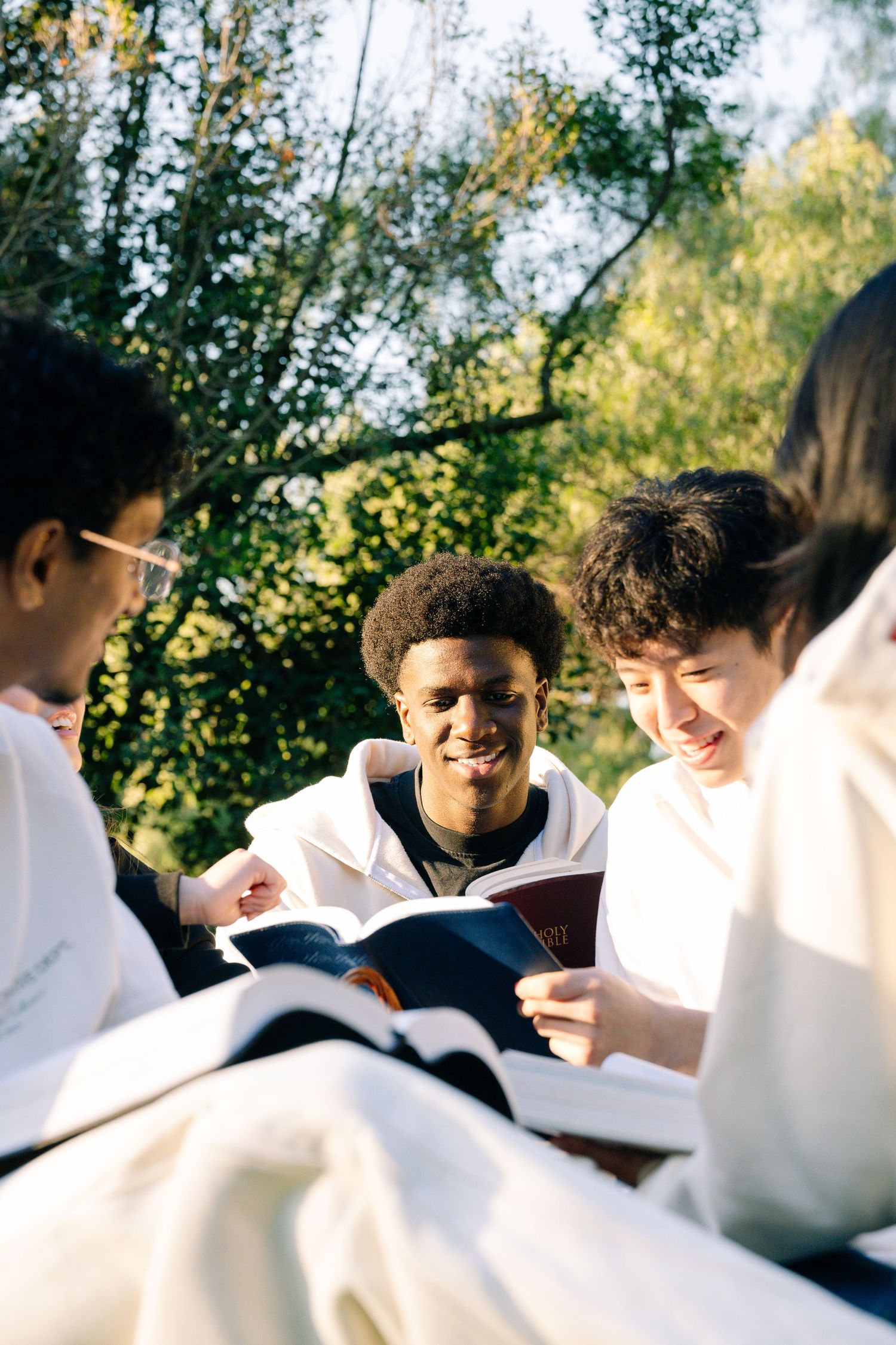 Diverse group of smiling young people reading books together outdoors in a sunlit park.