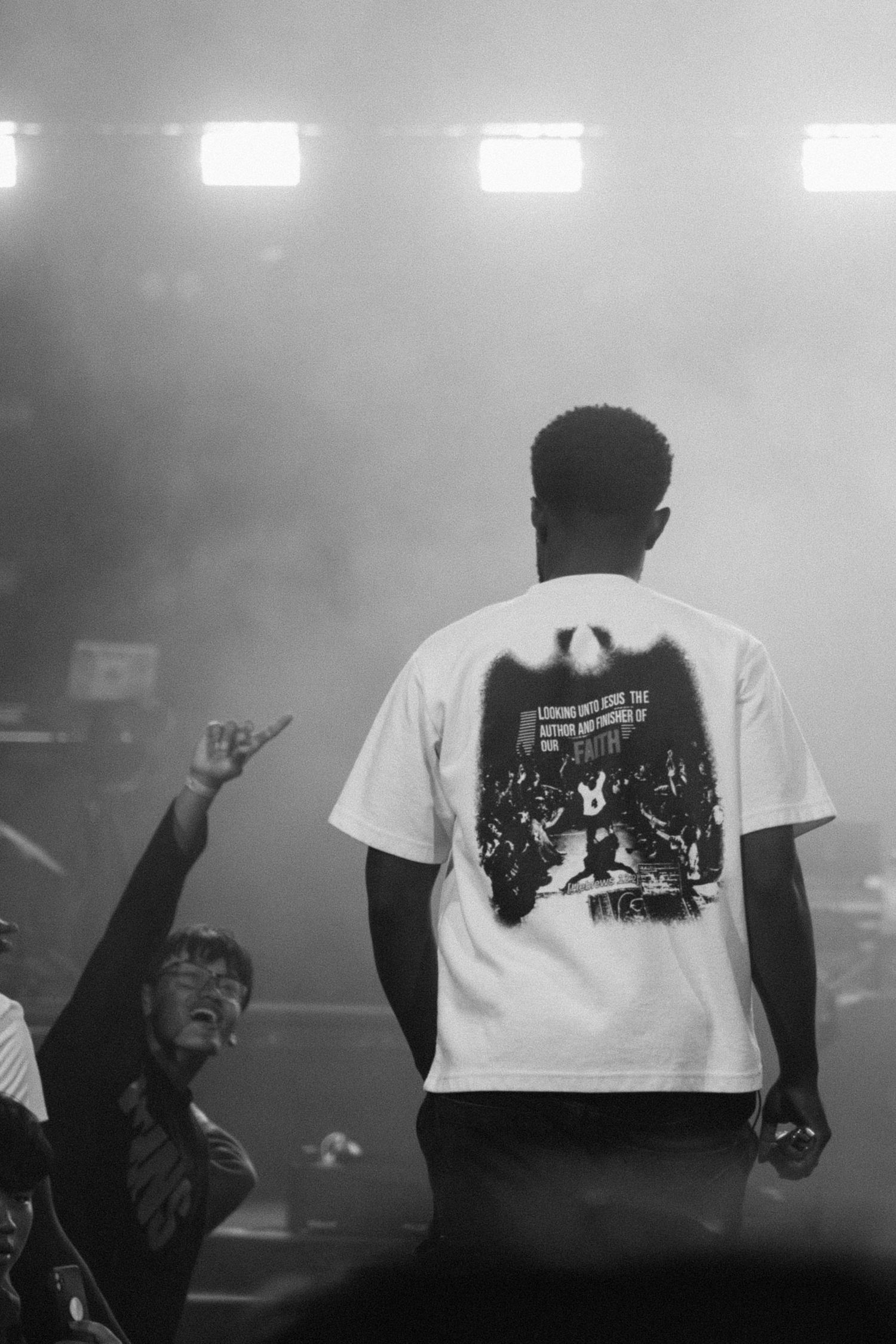 Black and white photo of a performer on stage in a graphic tee, with crowd members raising hands in smoky concert lighting.