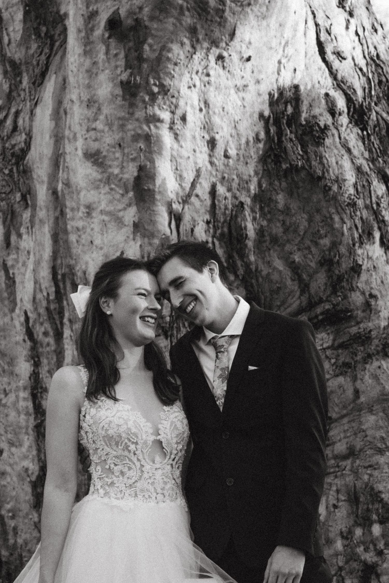 A laughing bride and groom lean together against a rocky backdrop in black and white.