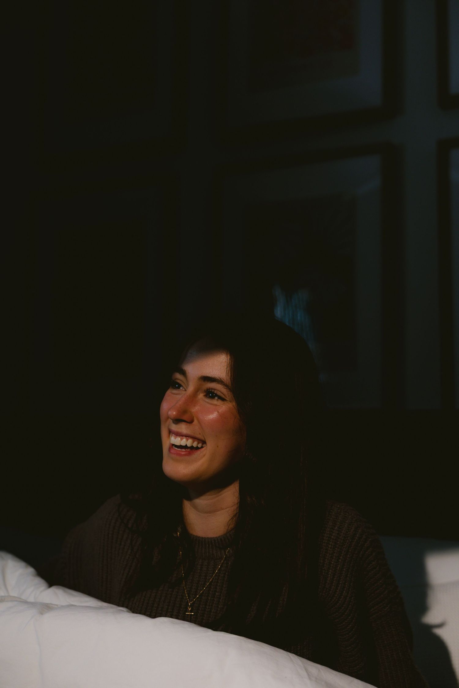 Smiling young woman with dark hair sitting in a dark room, illuminated by warm light.