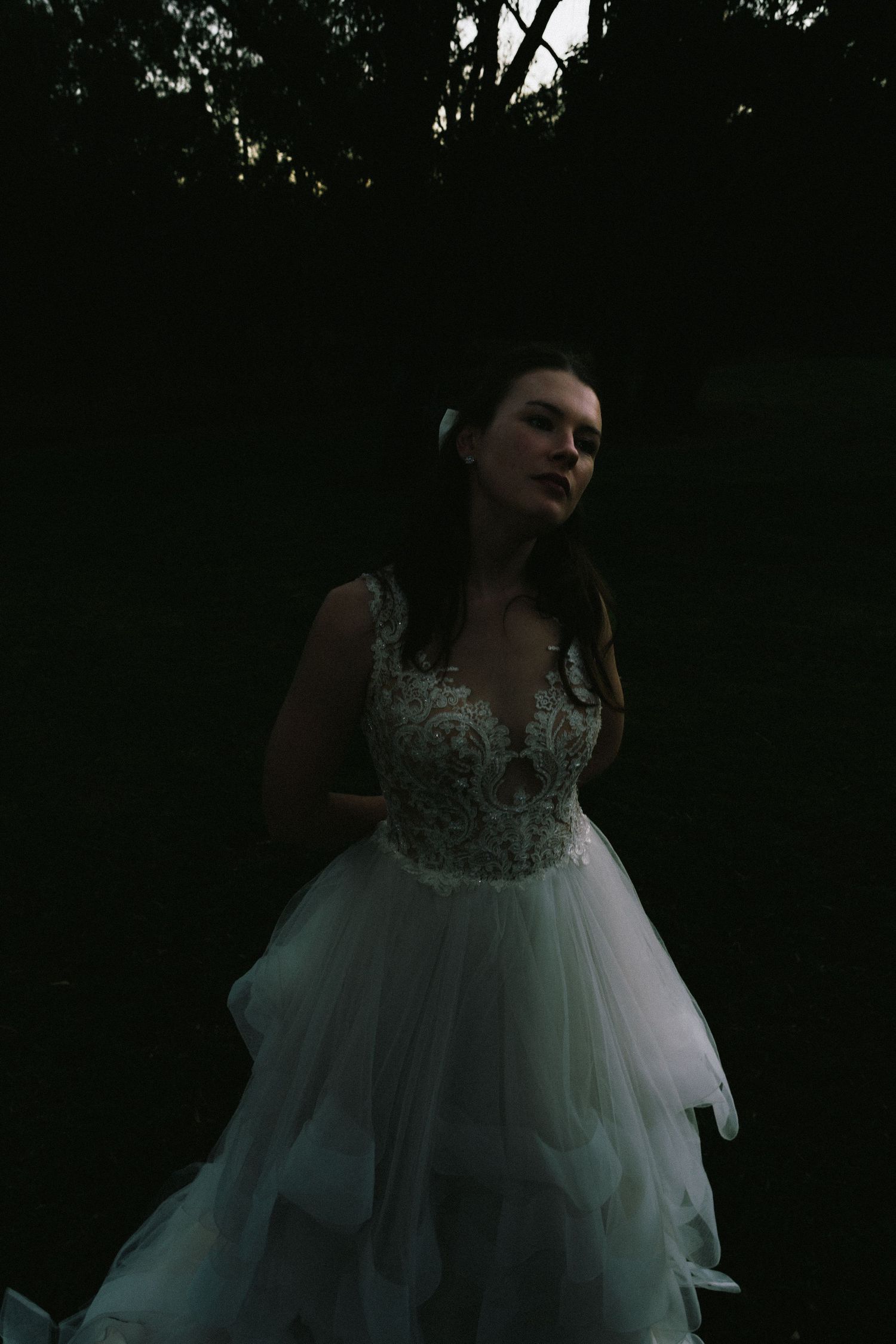 A bride in a lace and tulle wedding gown poses in moody dark forest lighting.