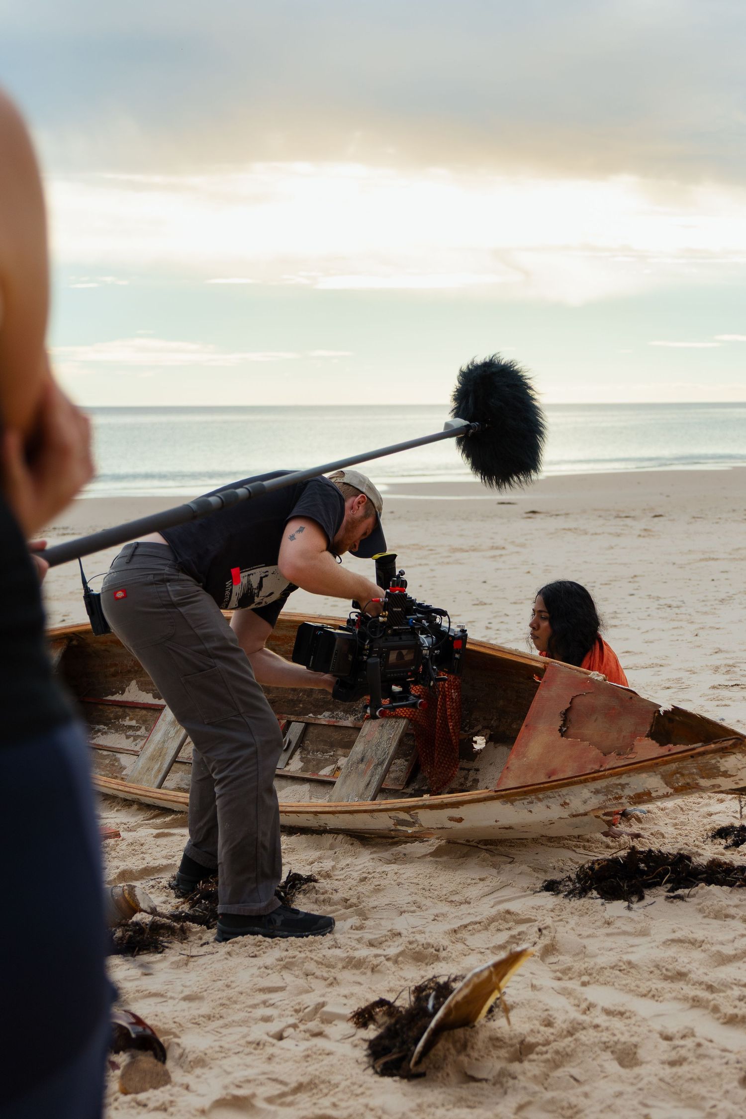 Cinematographer filming a person in an old wooden boat on a beach at sunset with boom mic overhead.