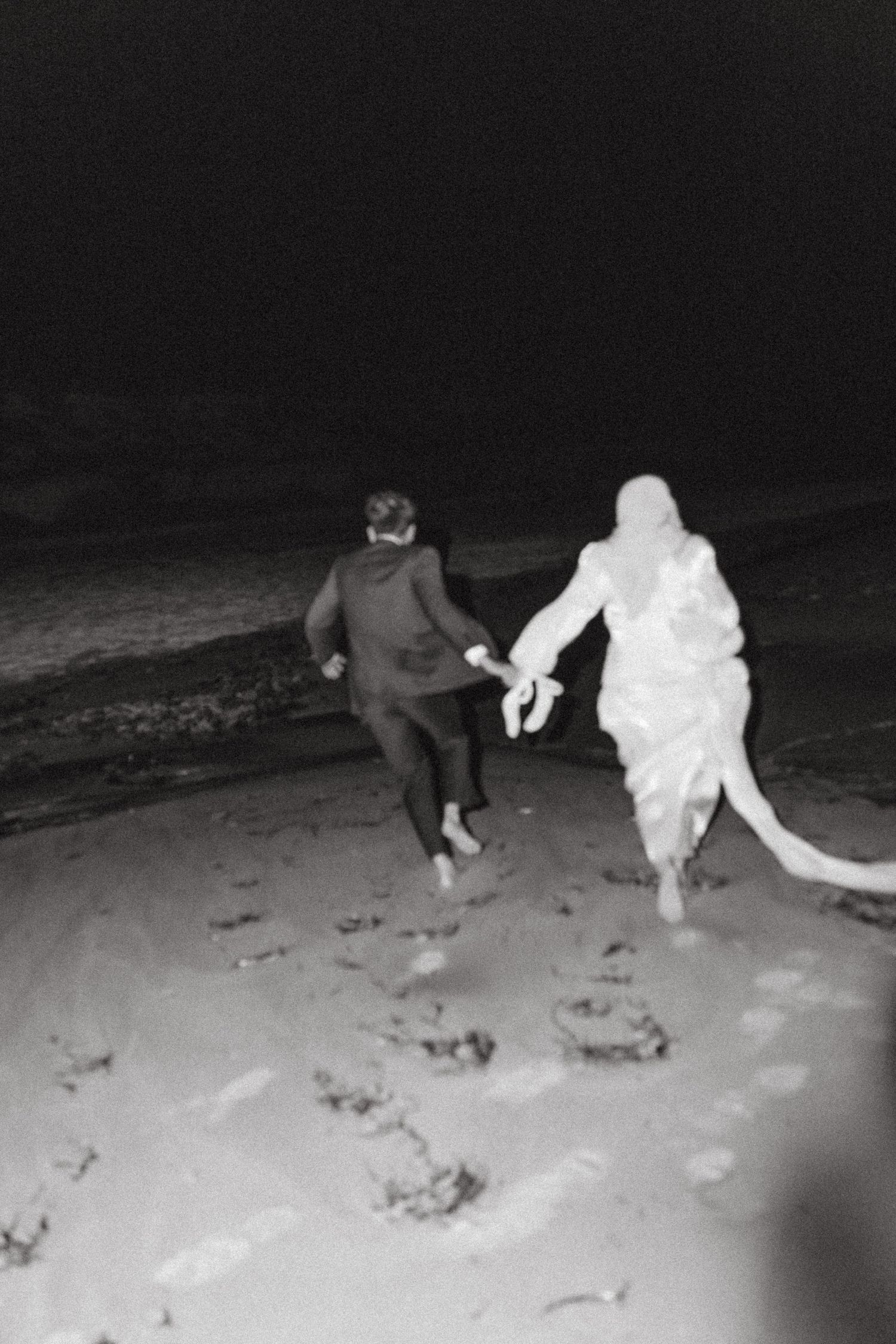 Black and white photo of a bride and groom running barefoot on a beach at night, holding hands.