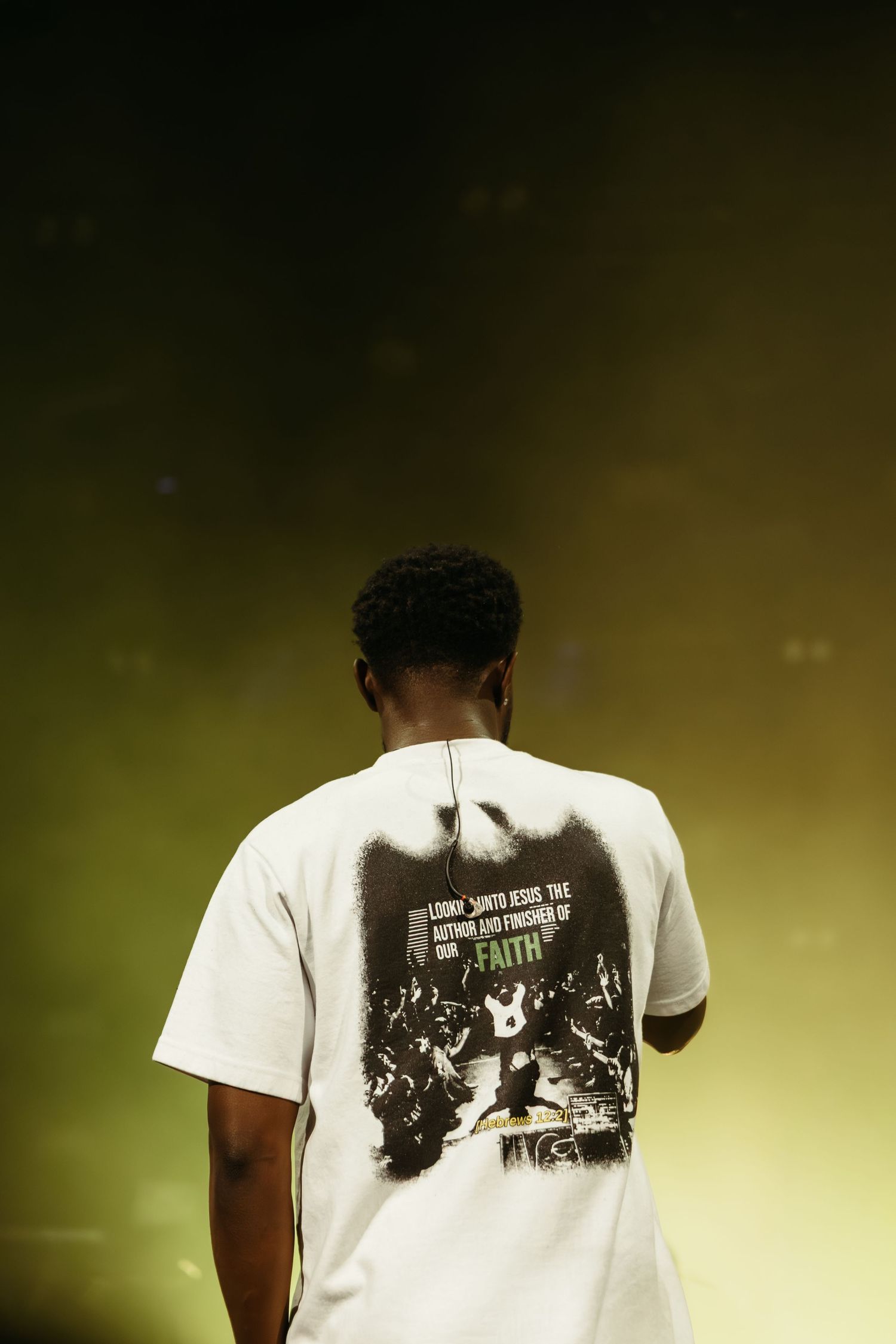 Person in white faith graphic tee stands on stage with back to camera under yellow-green spotlight.