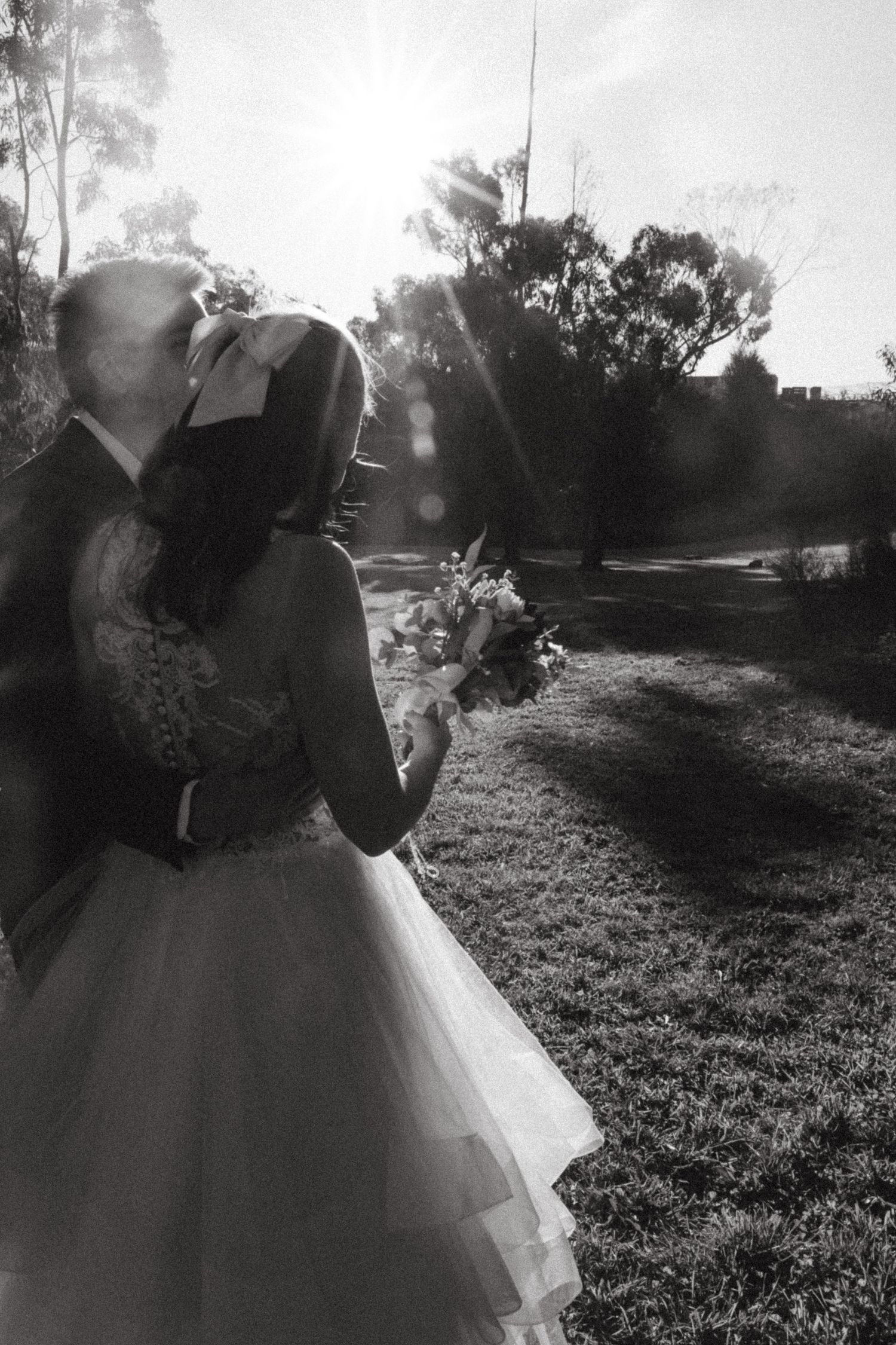 Black and white photo of newlyweds kissing outdoors with sun flare and bridal bouquet.
