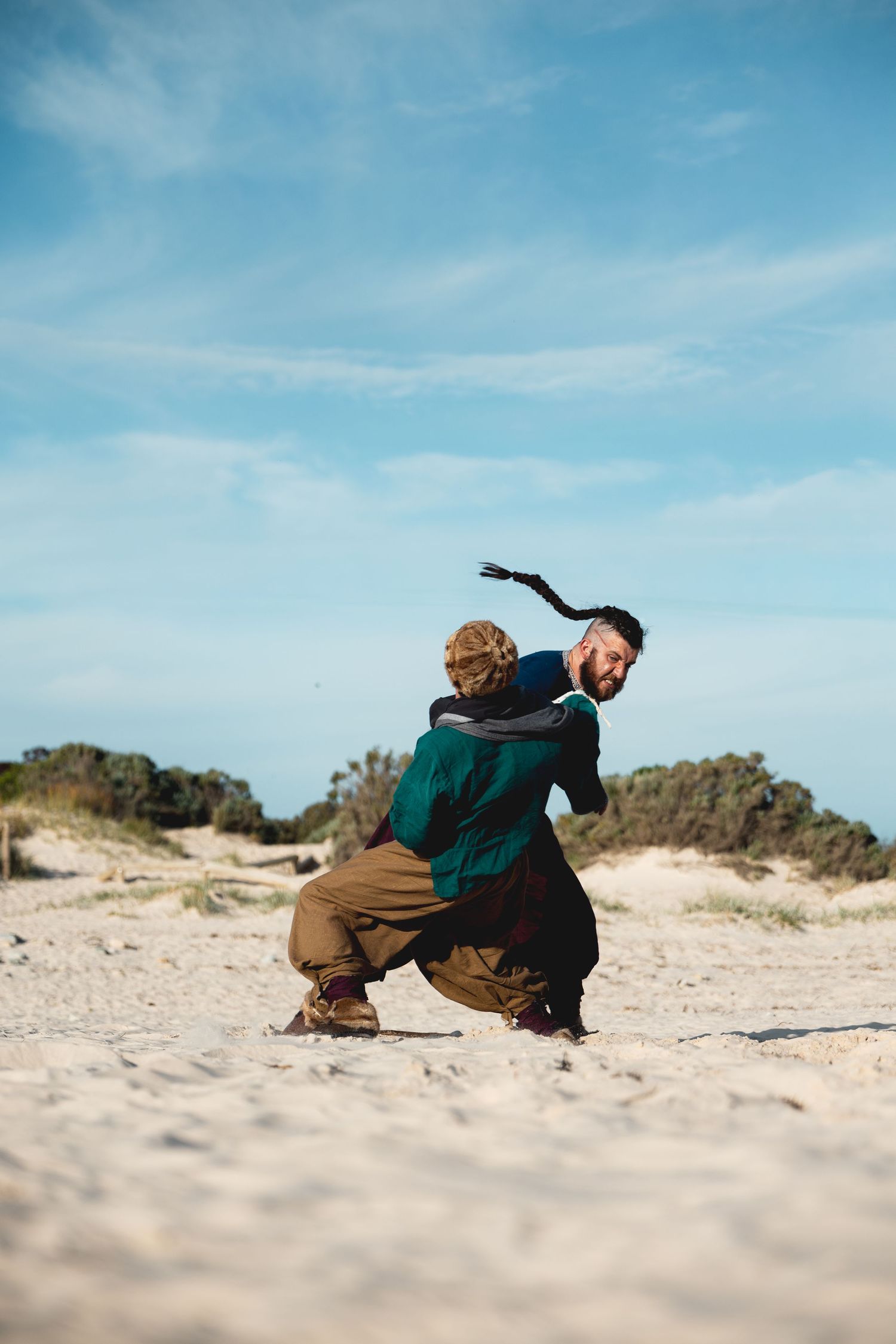 Two men in Viking costumes wrestling on sandy beach with green shrubs in background.