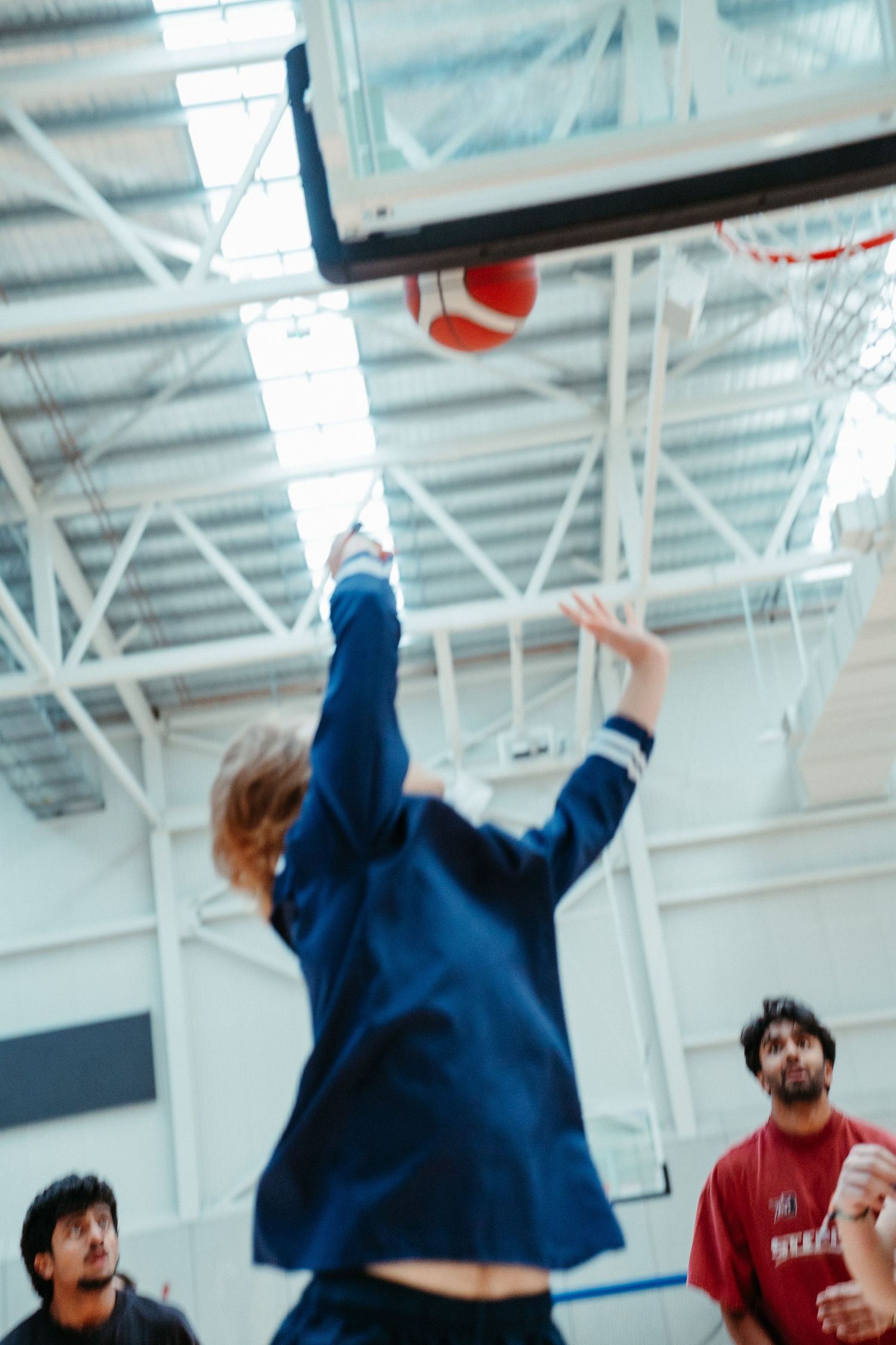 Player in blue jumpsuit leaping to shoot basketball in indoor gymnasium with spectators watching.