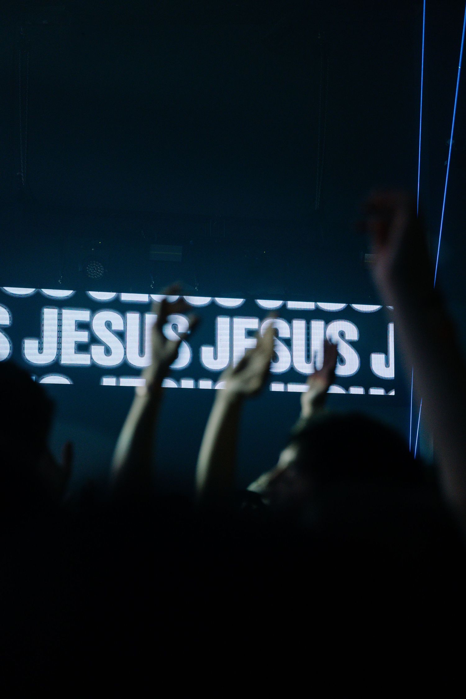 Crowd raises hands at a Christian worship concert with JESUS text displayed on a large LED screen in the background.