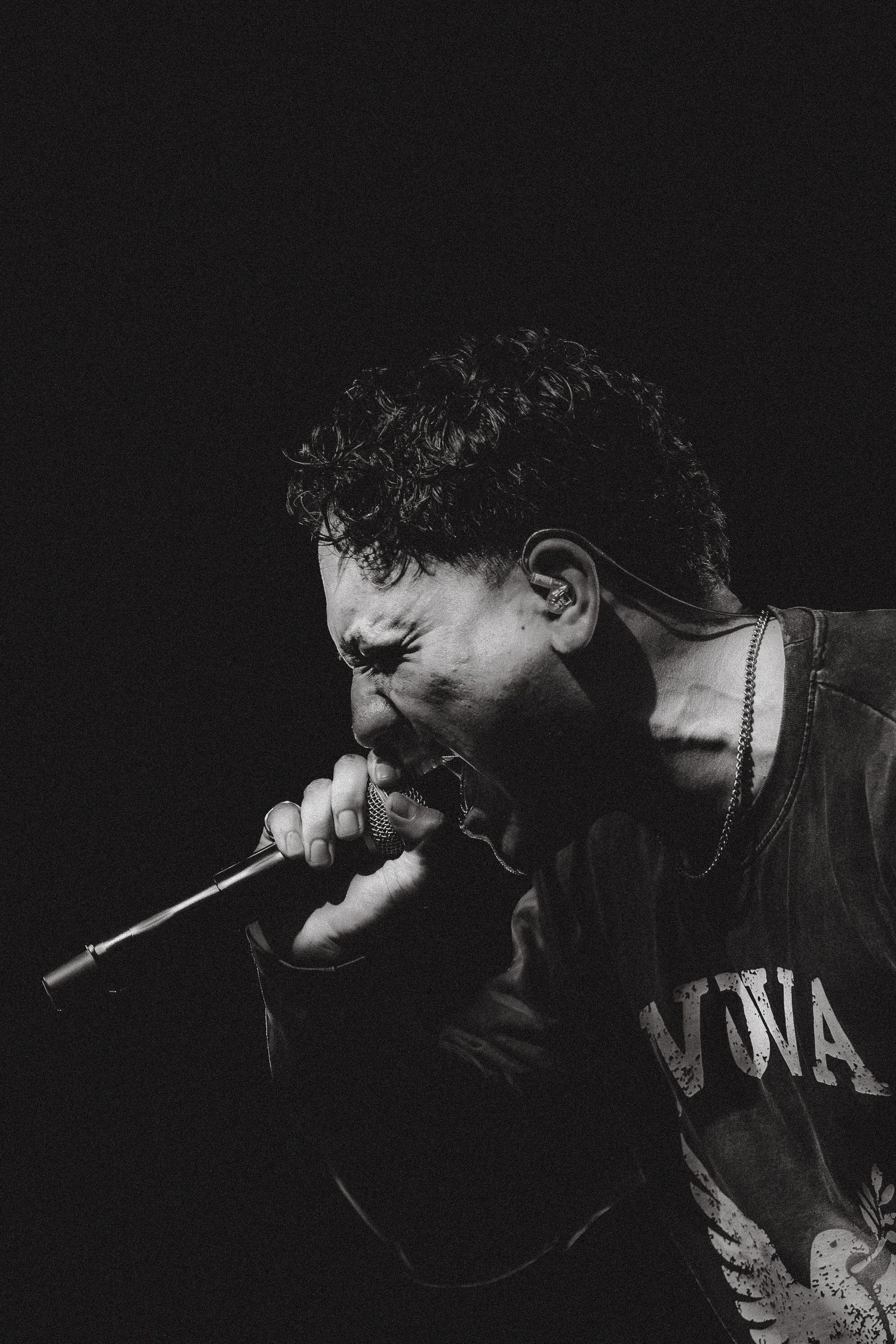 Black and white close-up of an intense male vocalist screaming into a microphone on stage.