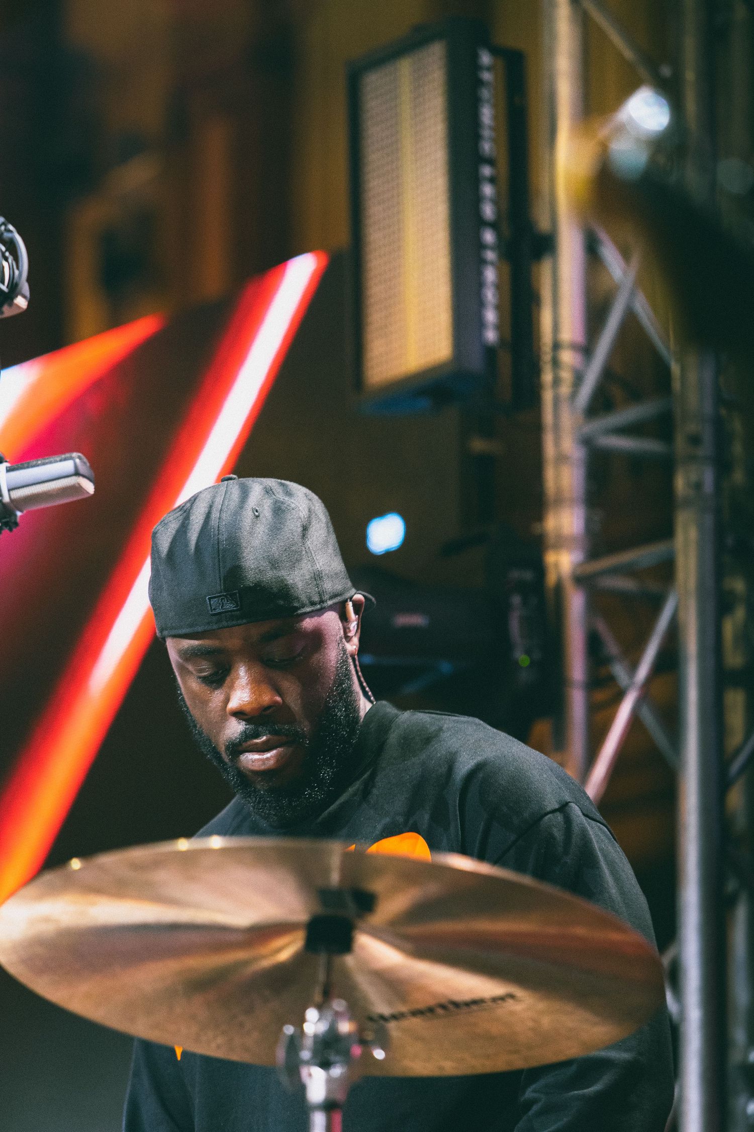 Male drummer performing on stage with cymbal under red stage lighting at live event.