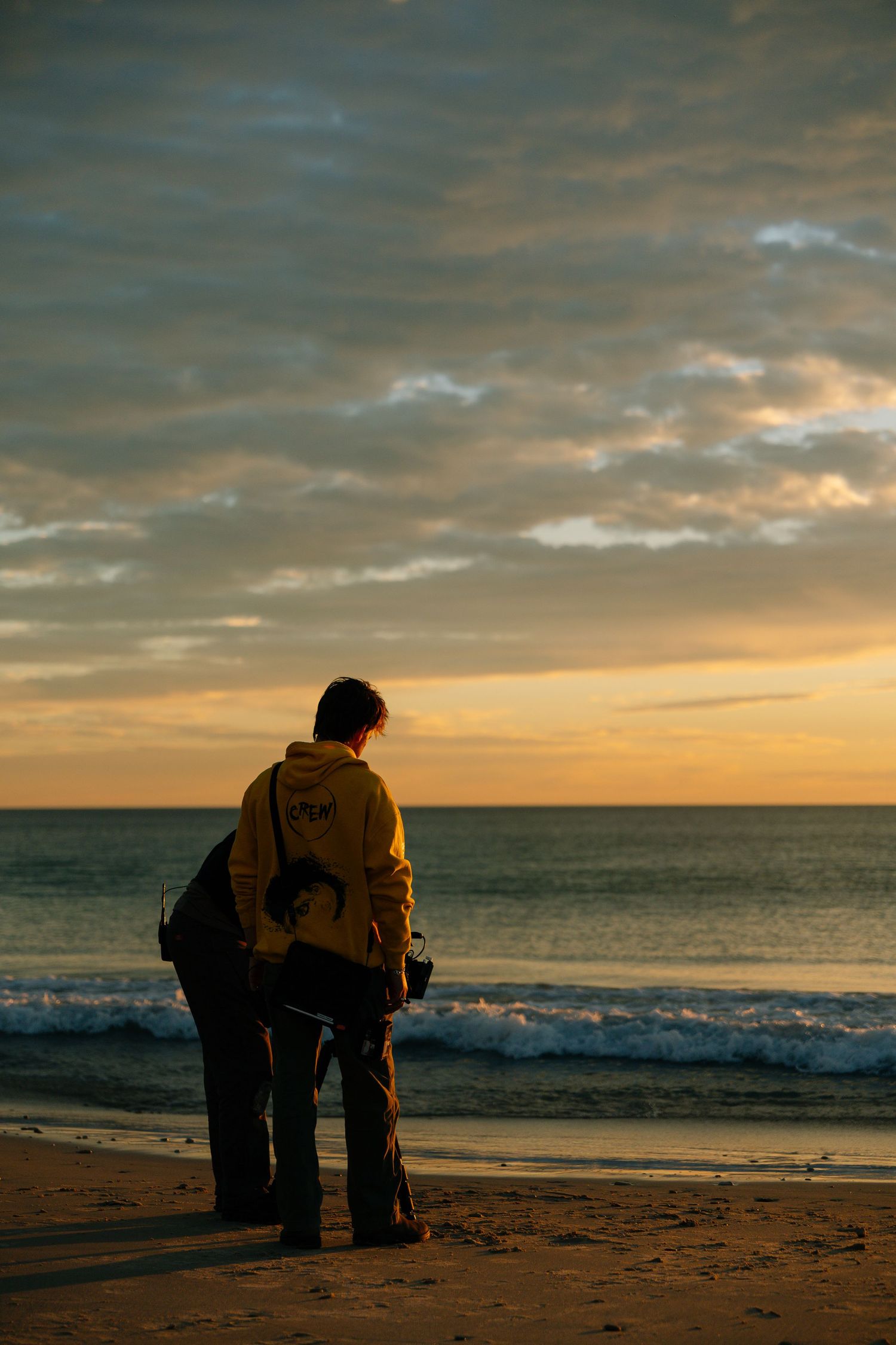 A lone filmmaker stands on a beach at sunset holding camera equipment by the ocean.