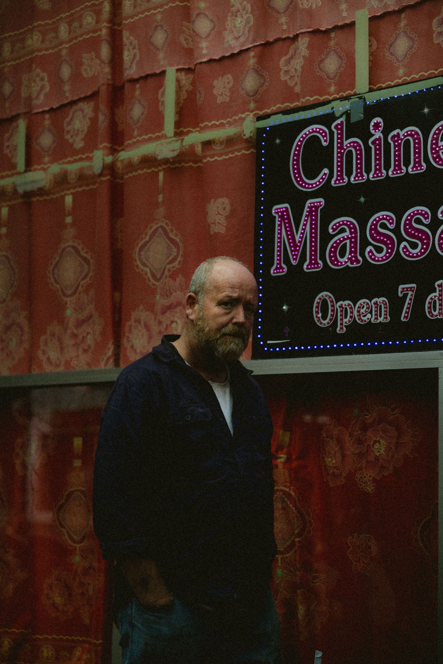 Bearded bald man standing outside a Chinese massage parlor with neon signage.