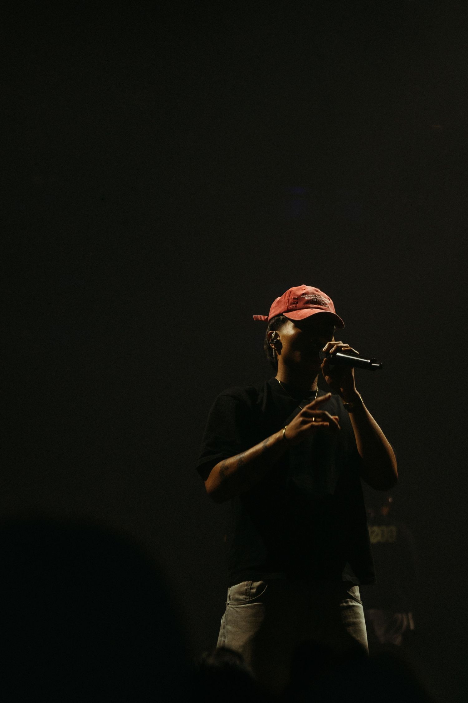 A performer in a red cap and black shirt holds a microphone on a dimly lit stage.