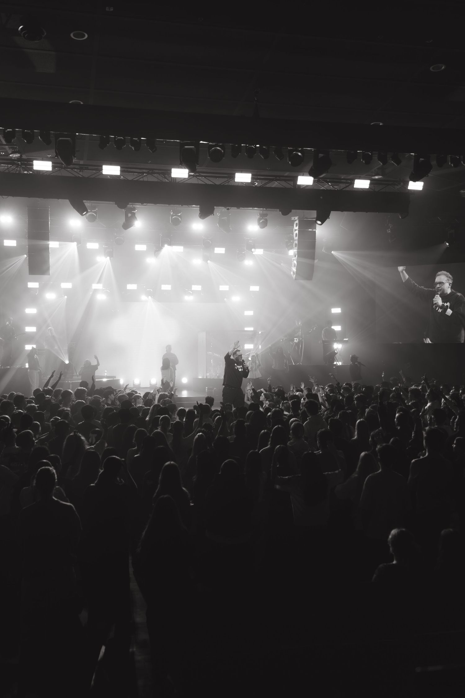 Black and white photo of a live concert with crowd silhouettes and bright stage lights illuminating performers on stage.
