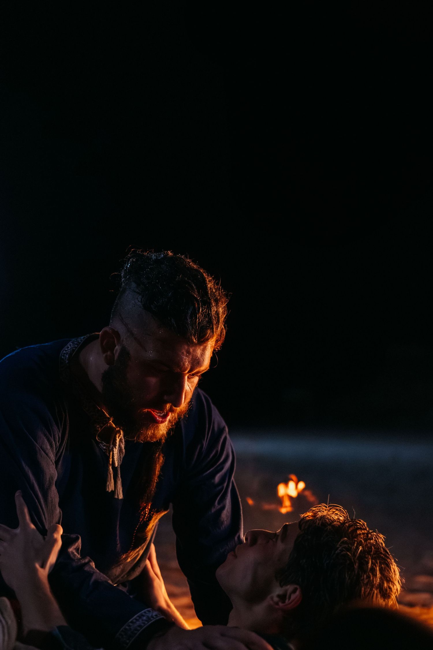 Dramatic firelit scene of a bearded man leaning over another person at night on a beach.