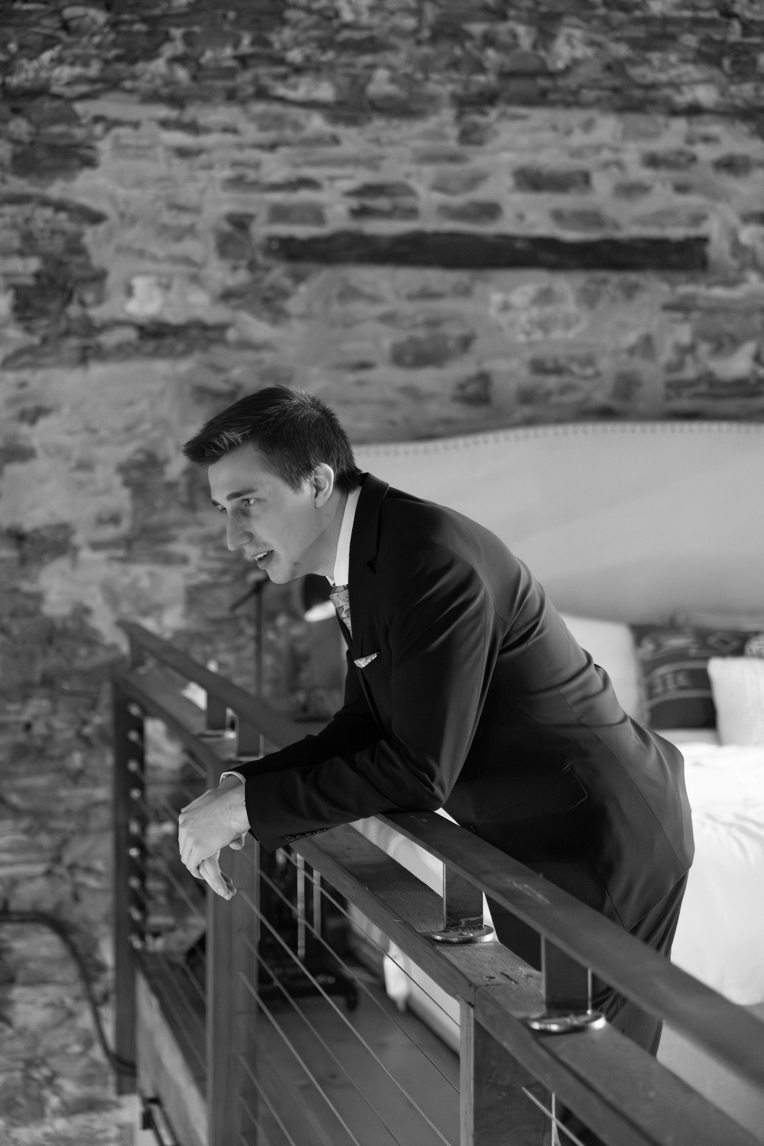 Black and white portrait of groom in tuxedo leaning on railing against rustic stone wall interior.