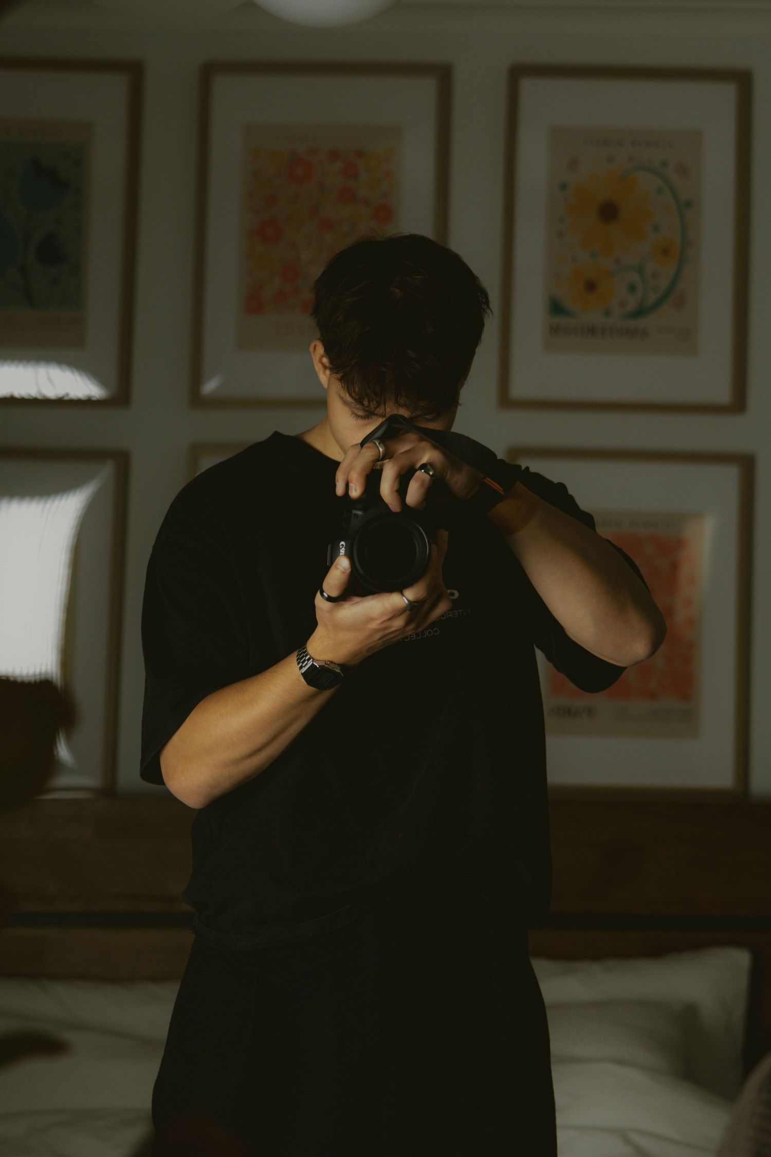 Man in black t-shirt holding a camera up to his face in a room with framed floral artwork.