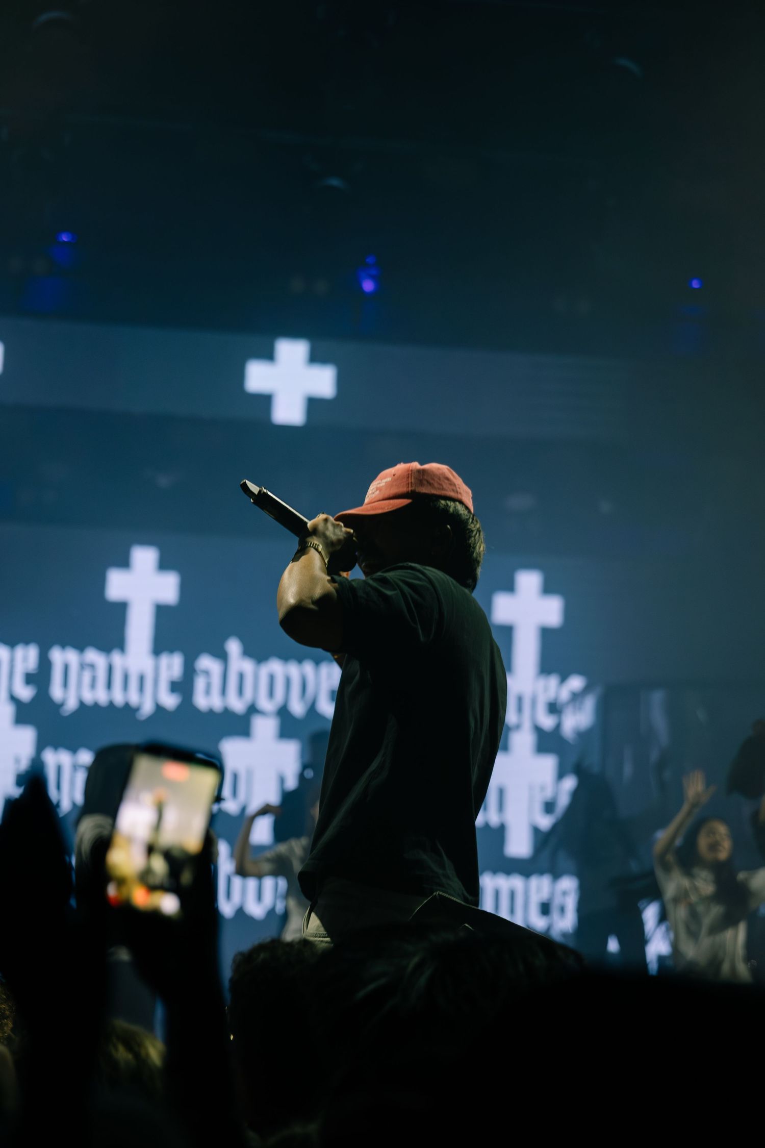 A rapper performs on stage with cross symbols and gothic text displayed on a screen behind them.