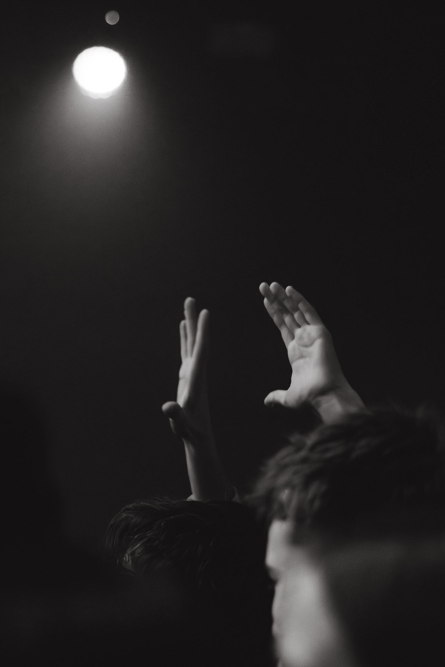 Black and white photo of hands raised in the air under a spotlight at a concert or worship event.