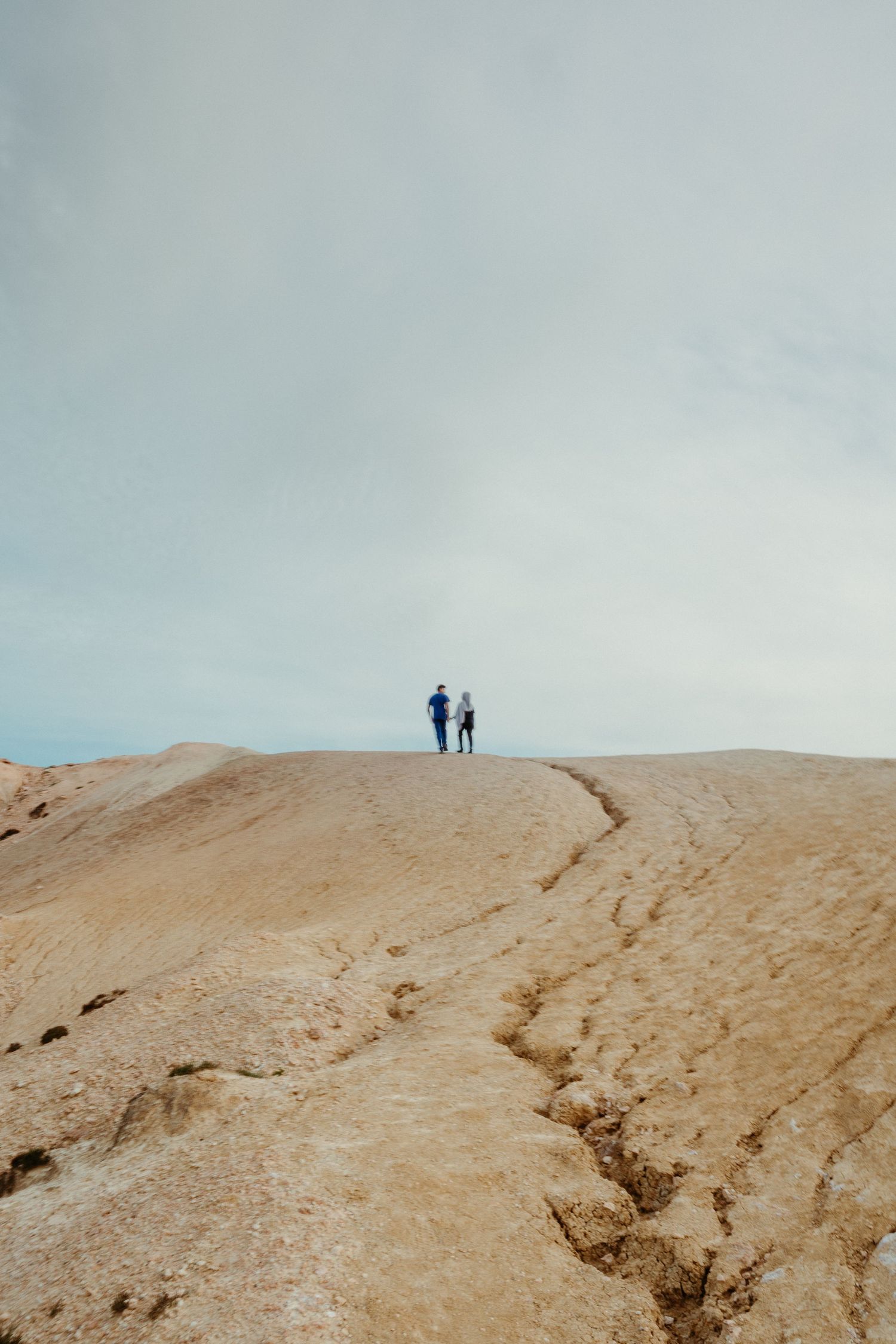 Two people stand atop a vast sandy rock formation under a cloudy sky in an arid desert landscape.