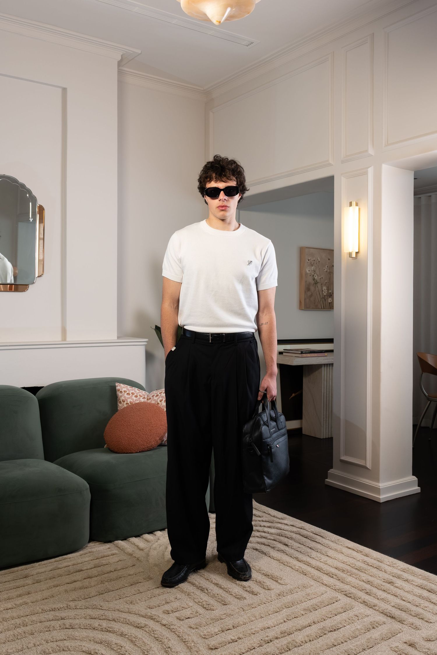 Young man in white t-shirt, black wide-leg trousers, sunglasses, holding bag, standing in elegant living room.