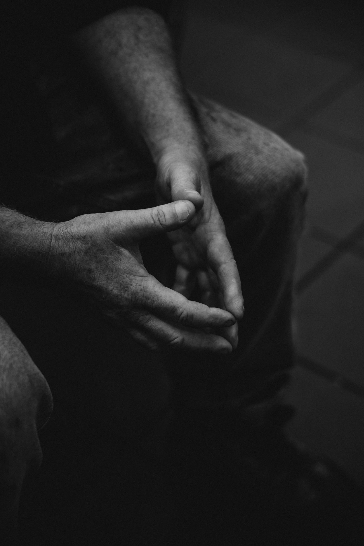 Black and white close-up of weathered male hands resting on knees in a dark setting.