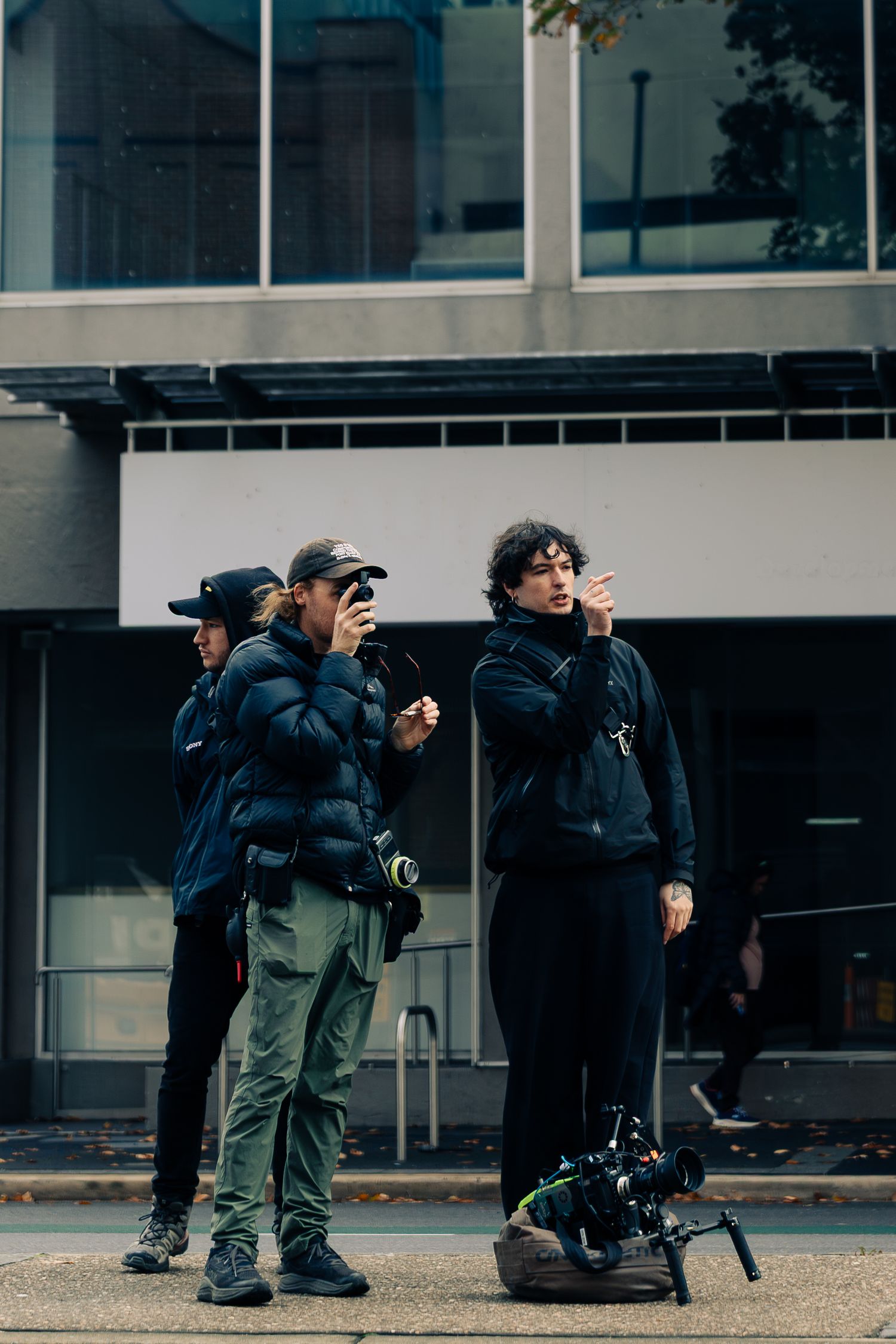 Film crew on urban street with camera equipment, two people reviewing shots on phones while a third observes.
