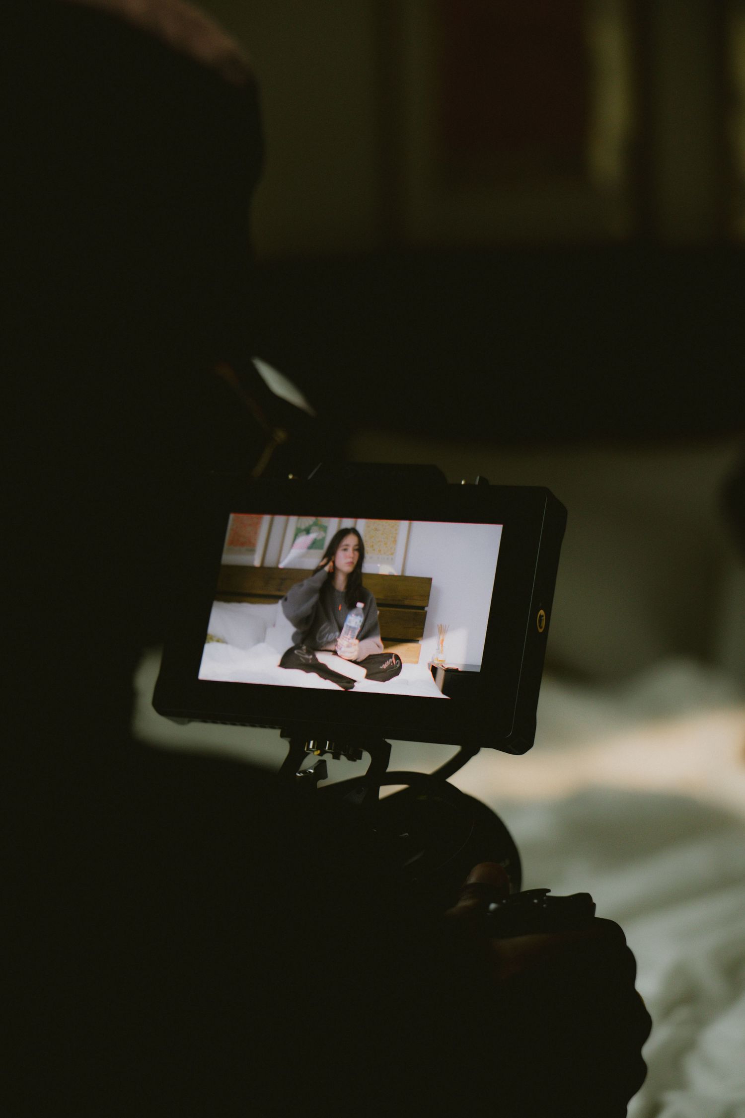 Filmmaker recording a woman sitting on a bed, visible on a camera monitor screen.