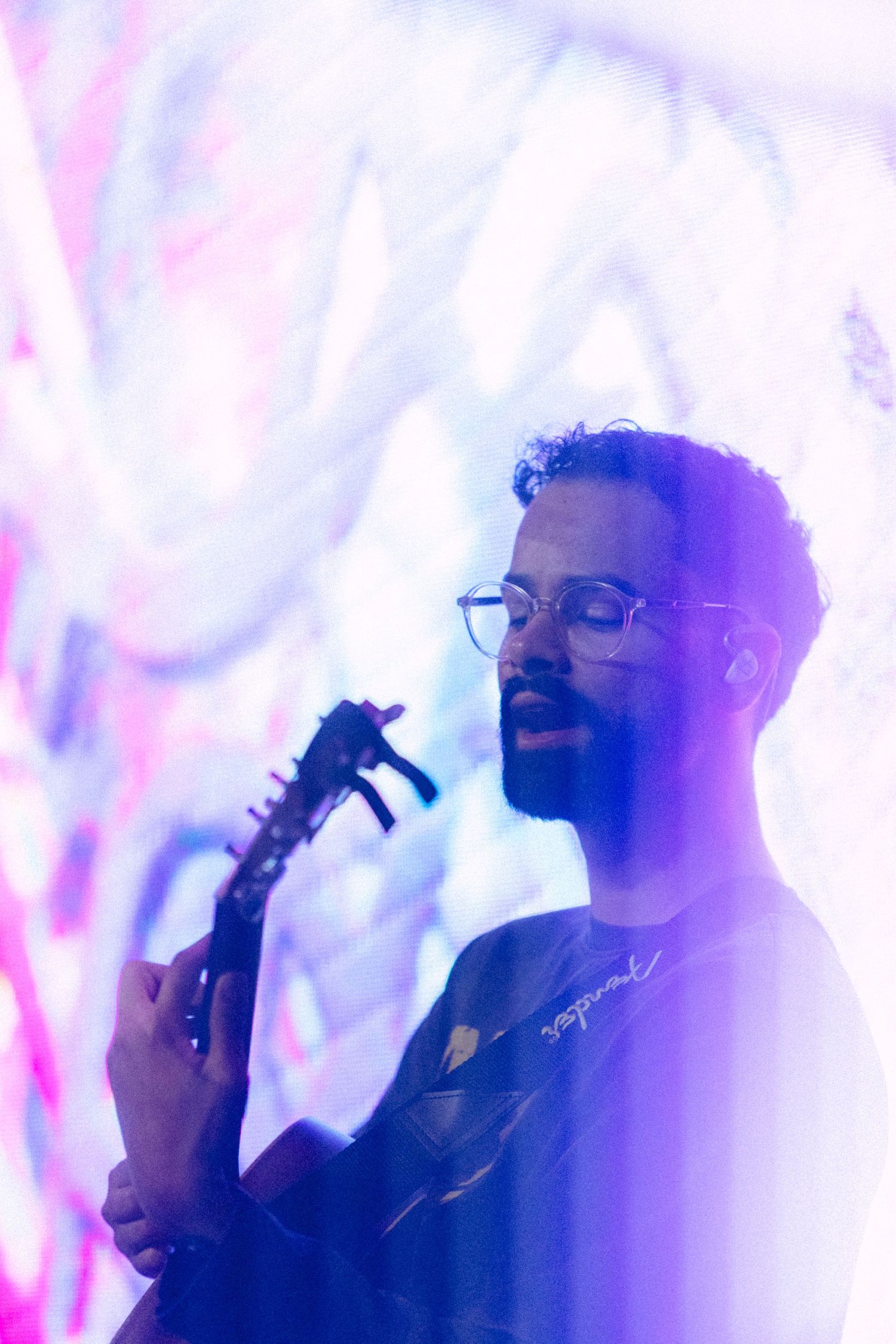 Bearded musician with glasses playing electric guitar bathed in purple and pink neon light backdrop.