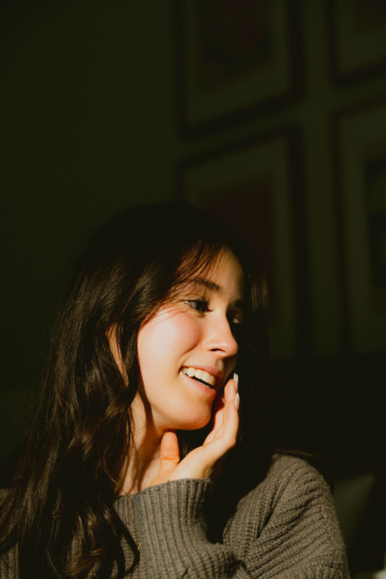 Young woman with long brown hair smiling softly, bathed in warm golden sunlight, wearing a gray knit sweater.