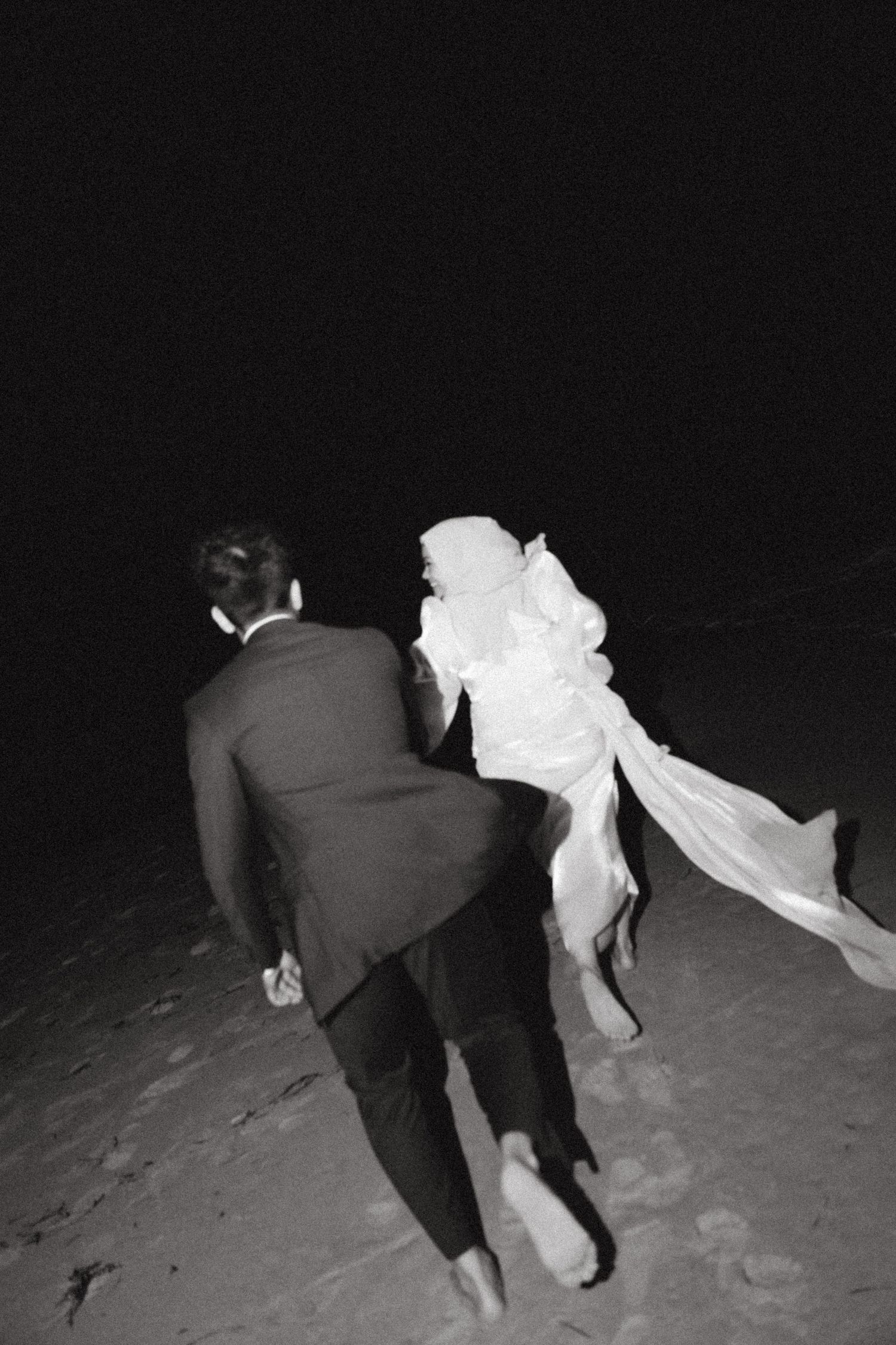 Black and white photo of a bride and groom walking barefoot on a beach at night, wedding veil flowing in the wind.