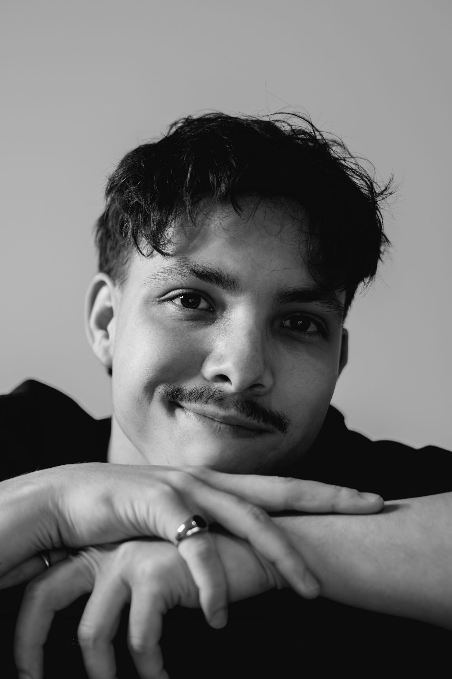 Black and white portrait of a young man with a mustache resting his chin on crossed hands, wearing a ring.
