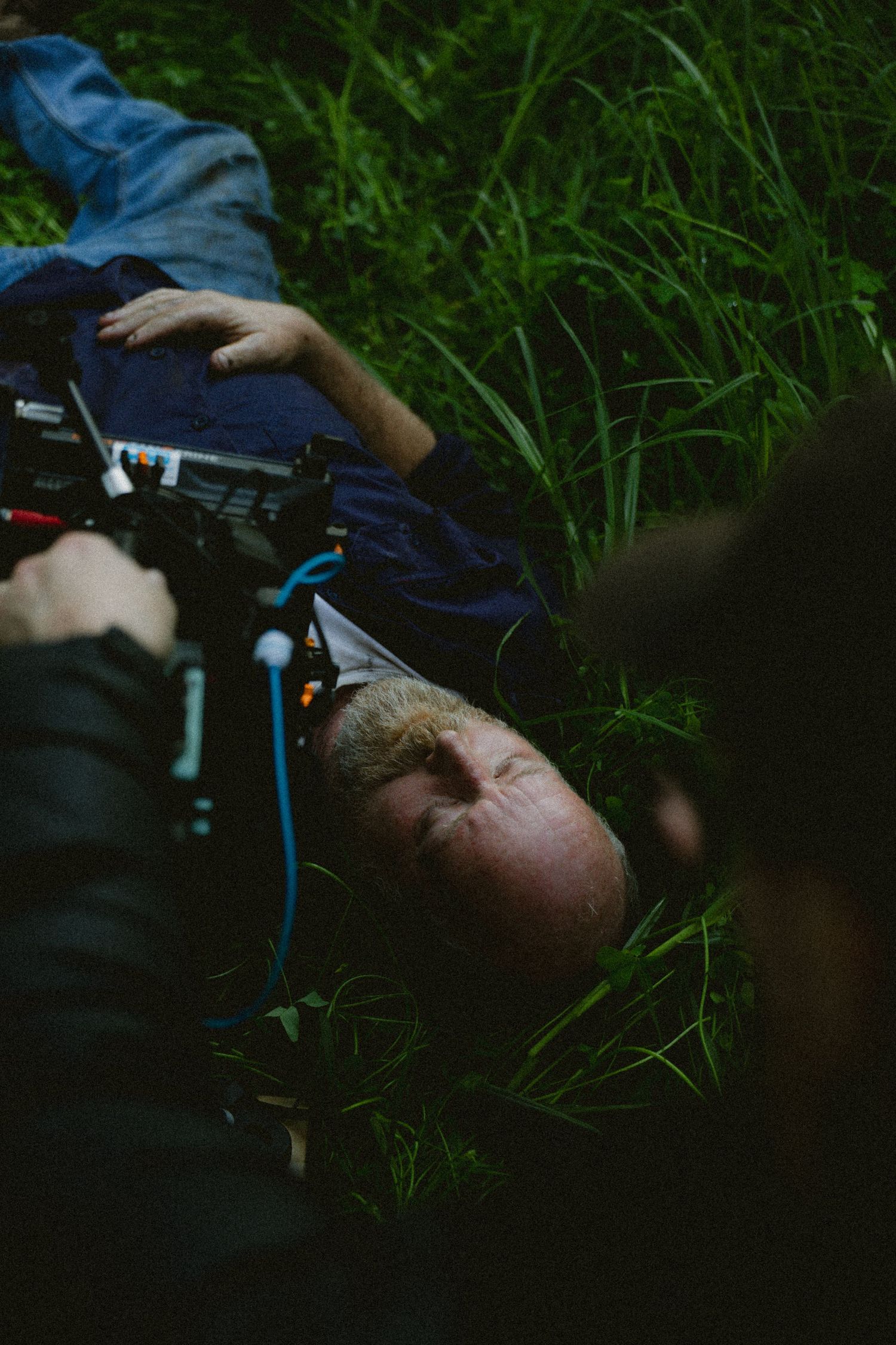 Cinematographer filming a bald man lying in tall green grass during an outdoor film shoot.