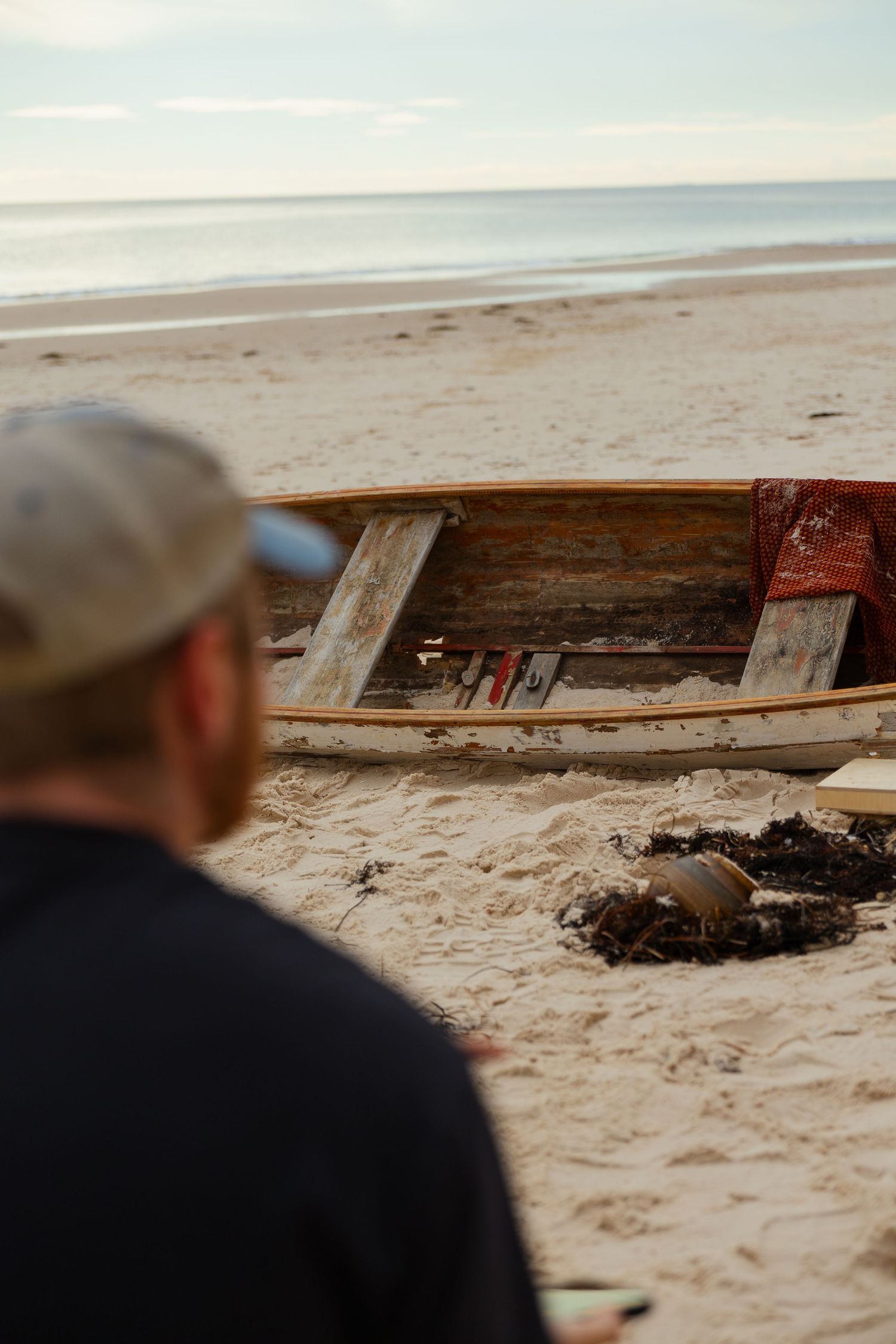 Man in a cap observing a weathered wooden boat on a sandy beach near the ocean.