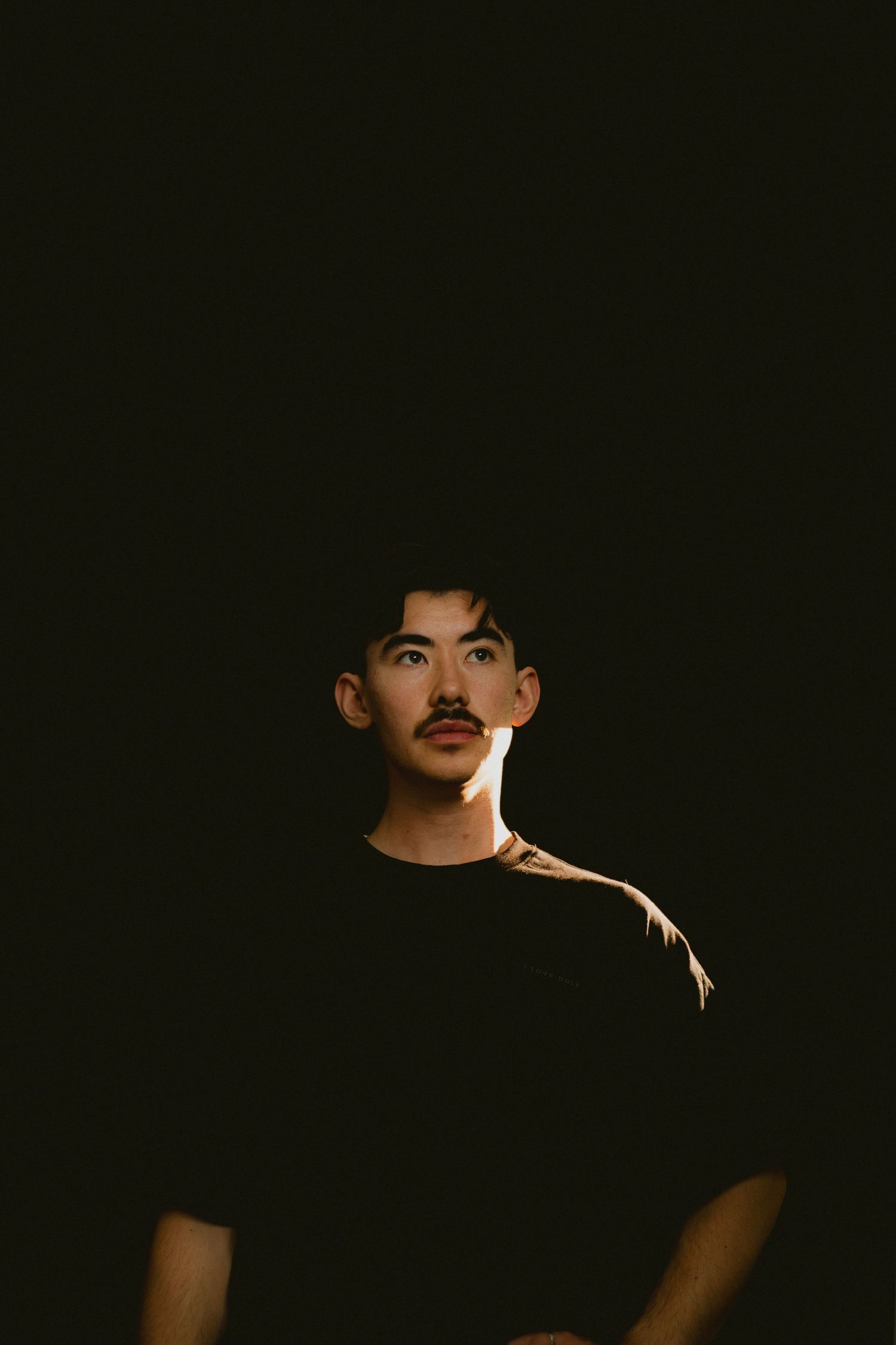 Young man with a mustache wearing a black t-shirt, dramatically lit against a dark background.