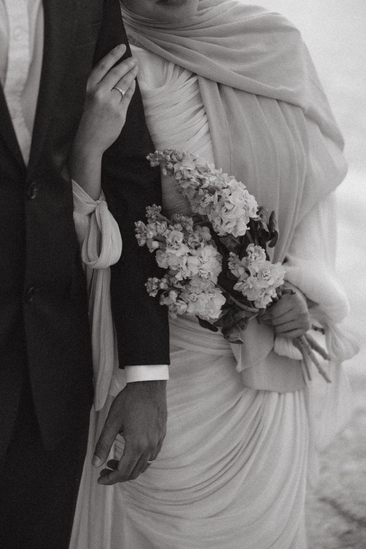 Black and white wedding photo of bride in draped gown holding floral bouquet beside groom in suit.