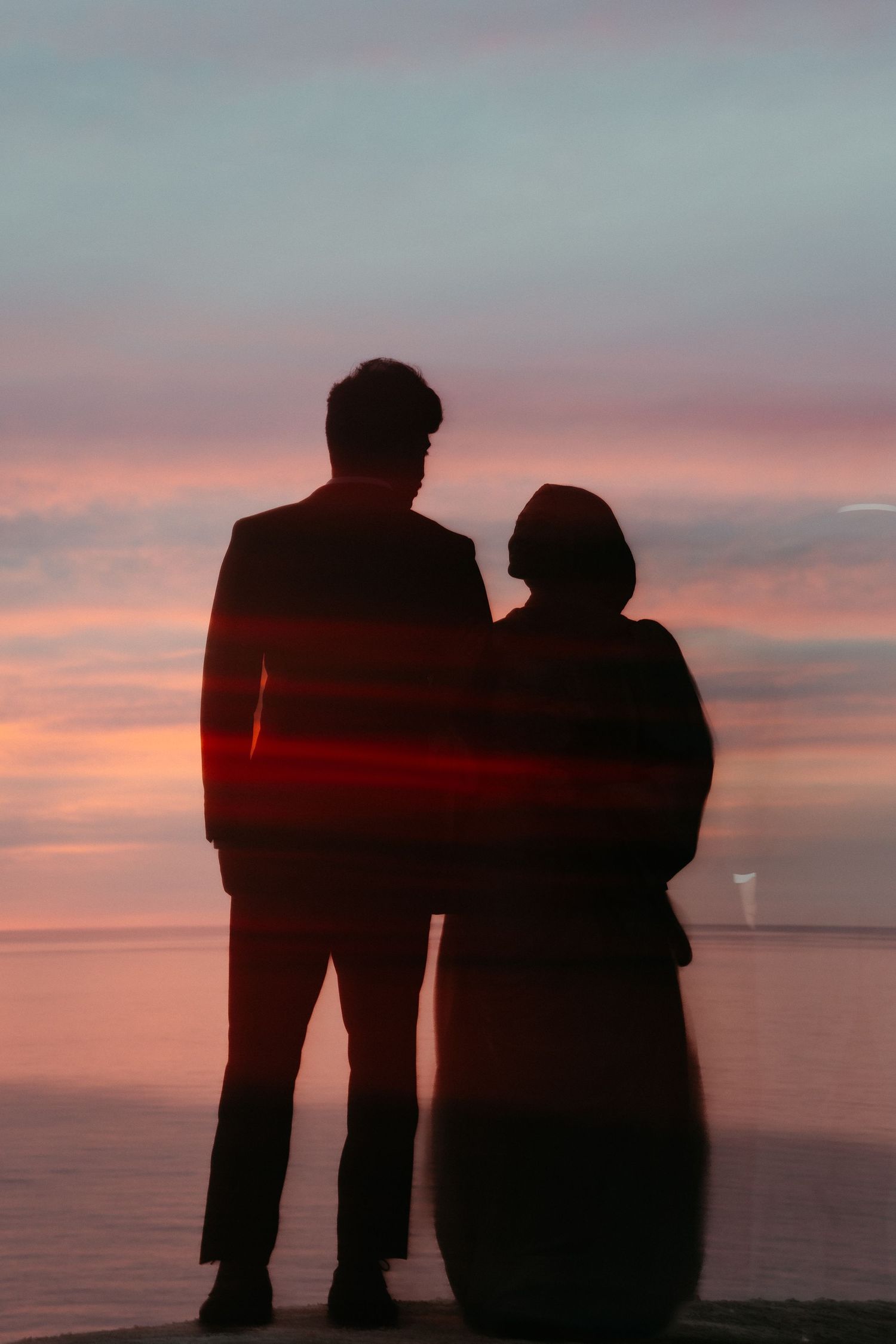 Silhouette of a romantic couple standing together watching a colorful pink and purple ocean sunset.