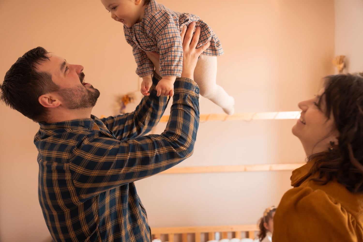 Un père soulève son bébé en l'air sous le regard souriant de la mère, moment de joie familiale lors d'une séance photo à domicile.