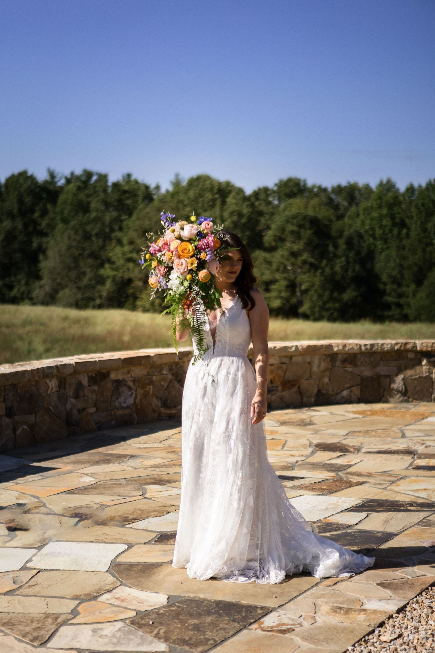 Bride in lace gown holds large colorful bouquet on stone terrace with forest backdrop.