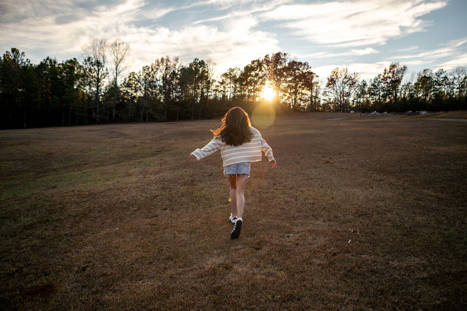 Young girl runs freely through an open field toward a stunning golden sunset in the countryside.