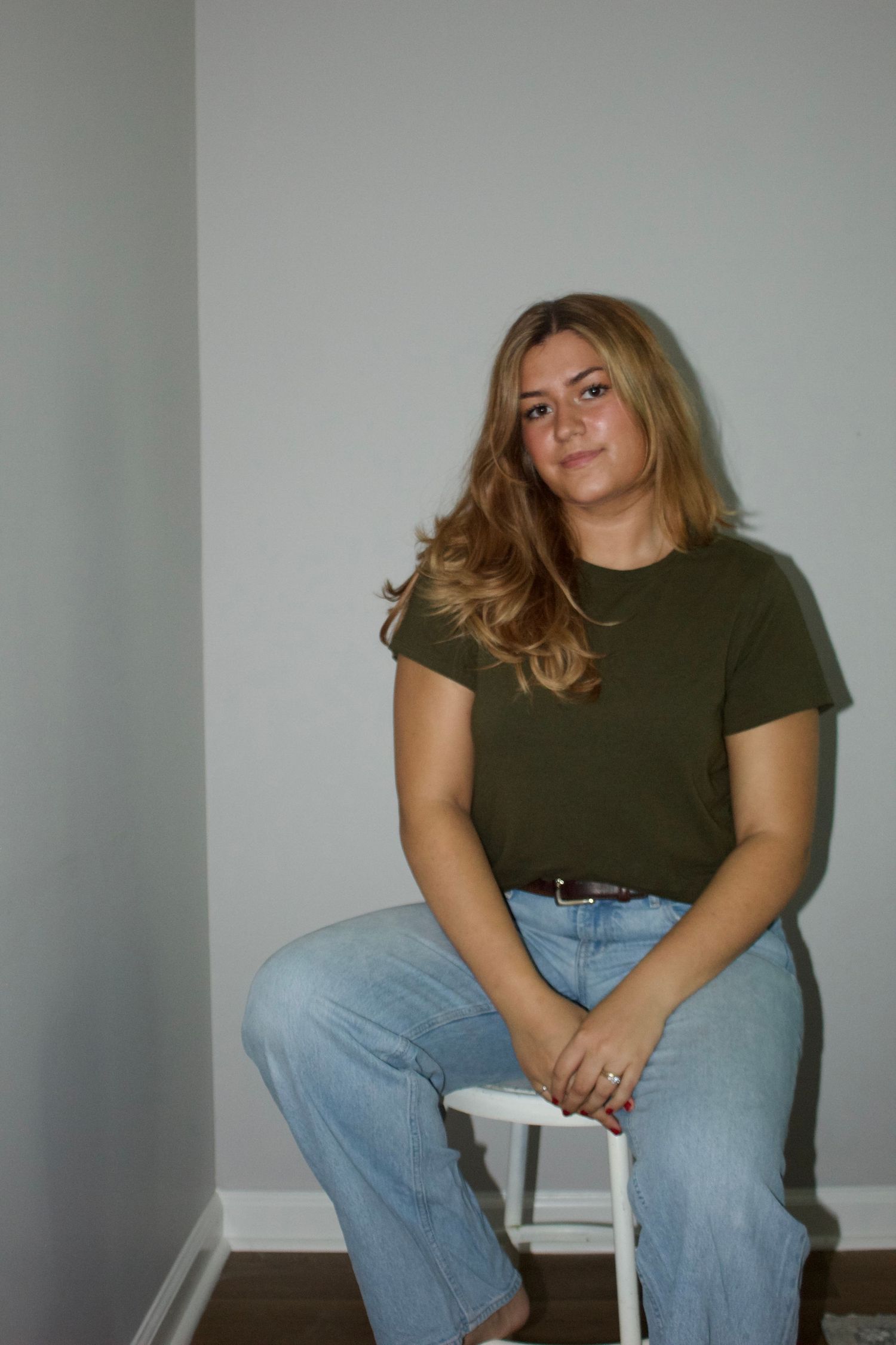 Young woman with wavy blonde hair sits on white stool wearing olive green t-shirt and light blue jeans.