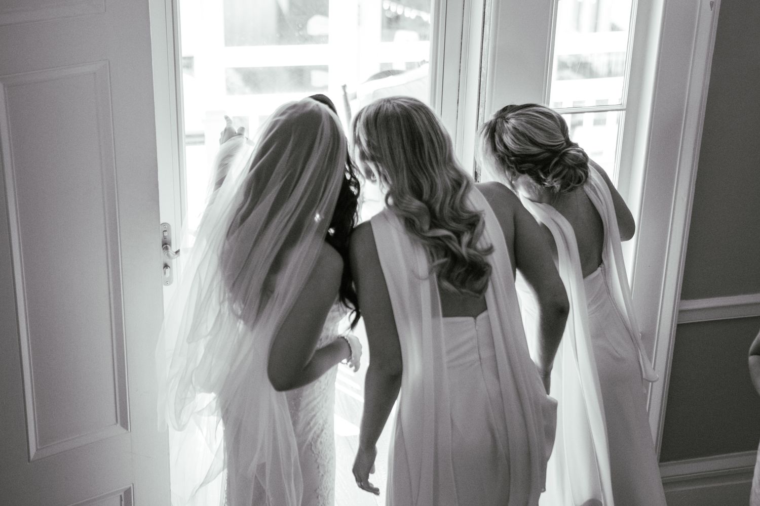 Bride with veil and two bridesmaids in white gowns seen from behind standing at bright doorway, black and white.