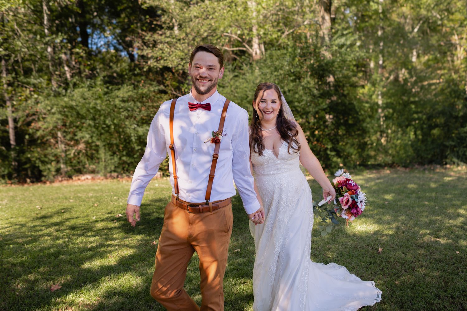 Happy groom in suspenders and bow tie walks with smiling bride holding colorful bouquet on garden lawn.