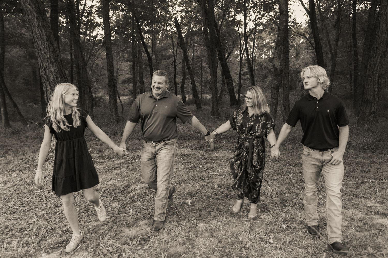 Black and white photo of a family of four holding hands walking through a wooded area.