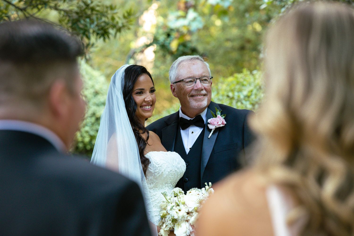 Radiant bride in strapless gown smiles with older gentleman in tuxedo during outdoor garden wedding ceremony.
