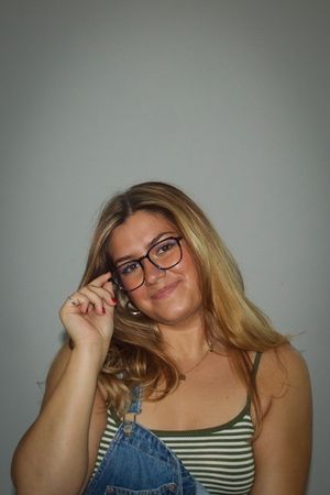 Blonde woman in glasses and denim overalls adjusting her frames while smiling at camera.