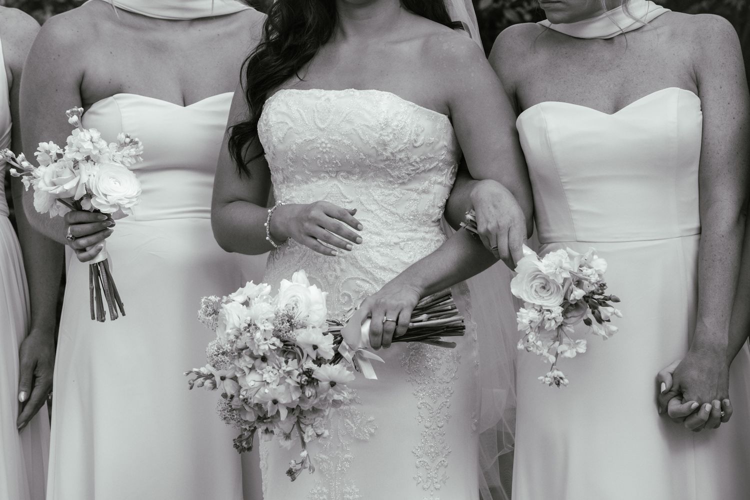 Bride in lace strapless gown stands with bridesmaids in white dresses holding floral bouquets, black and white photo.
