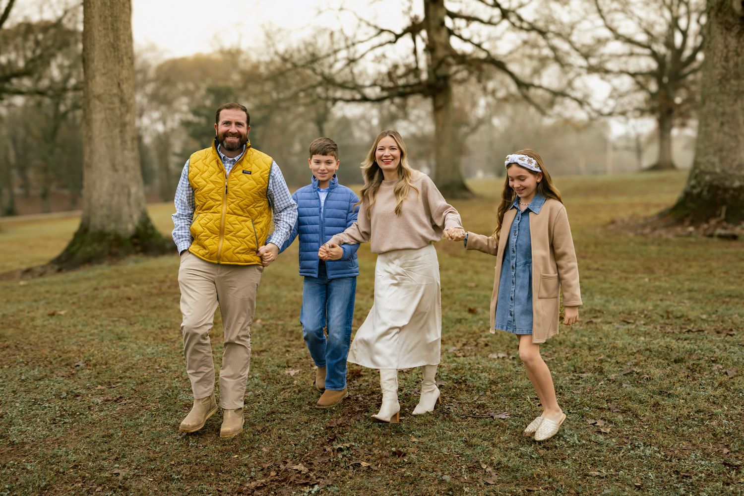 Smiling family of four walks together through autumn park during outdoor family portrait session.